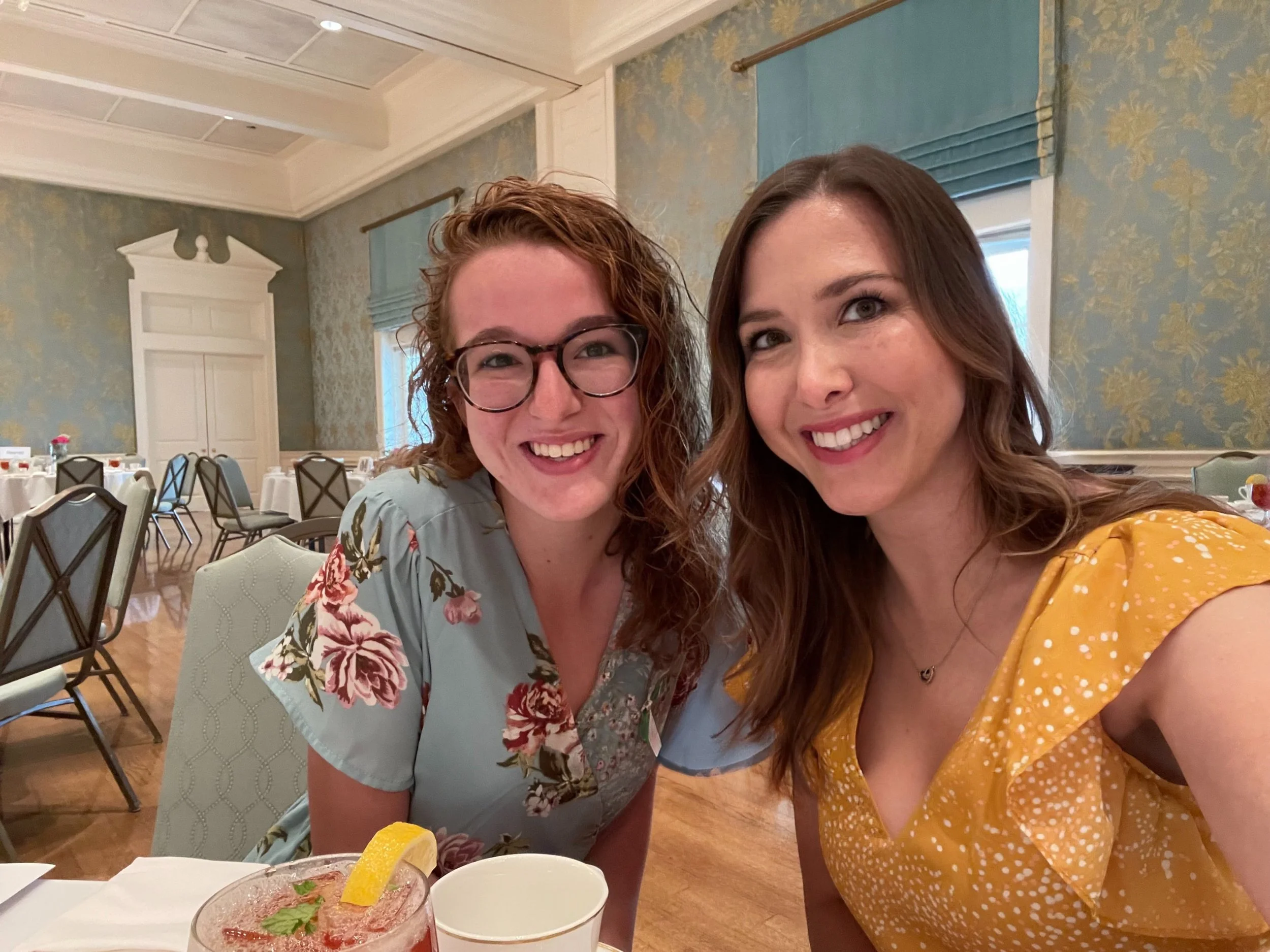 Two women sitting at a table in a decorated dining room, smiling at the camera. One woman has curly red hair and glasses, wearing a floral blouse. The other has long brown hair, wearing a yellow dress. There are plates, cups, and a drink with a lemon