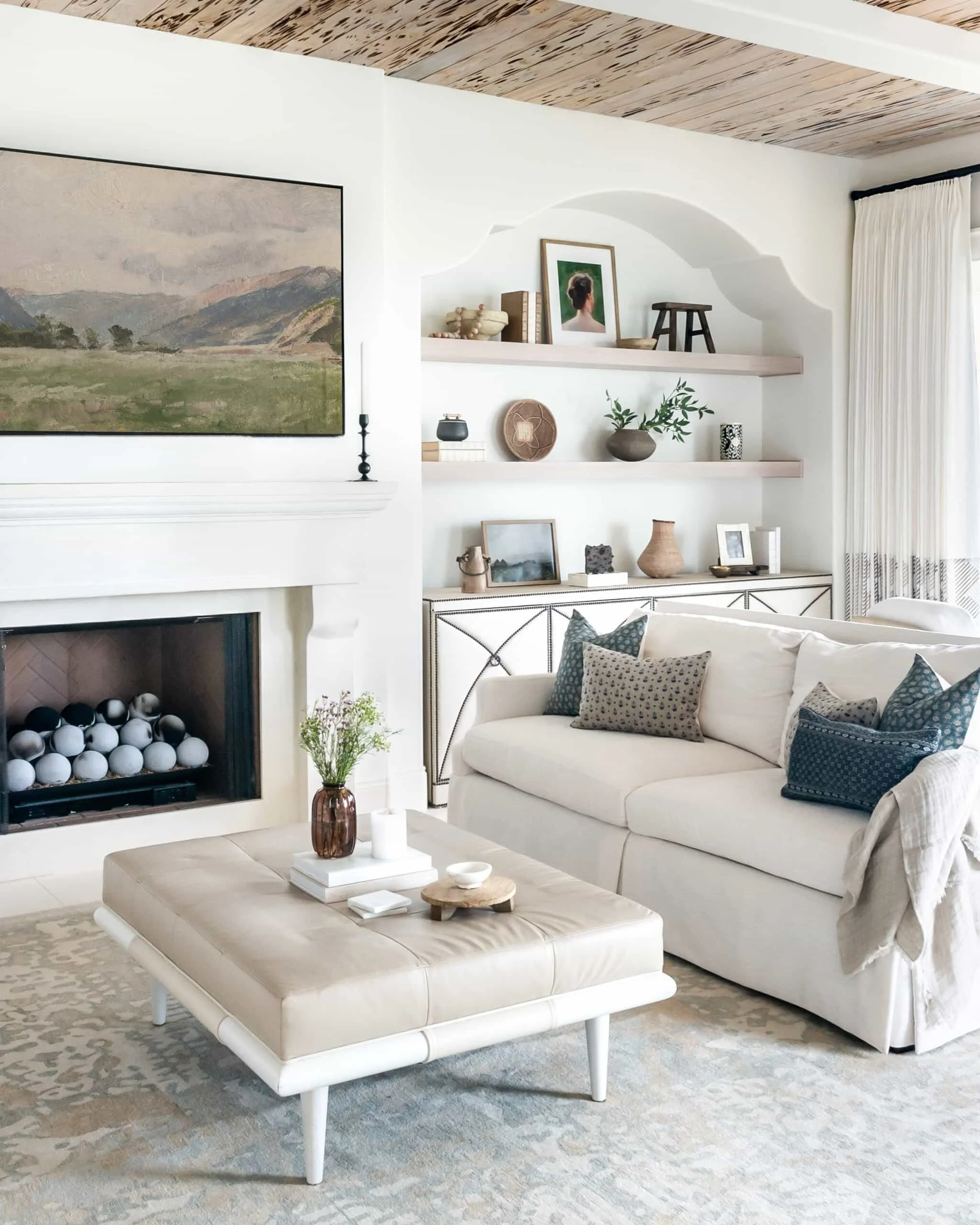 Living room with a beige ottoman and a white sofa with patterned pillows, a fireplace with decor, a shelf with various decorative items and picture frames, a vase with flowers, and a large landscape painting above the fireplace.