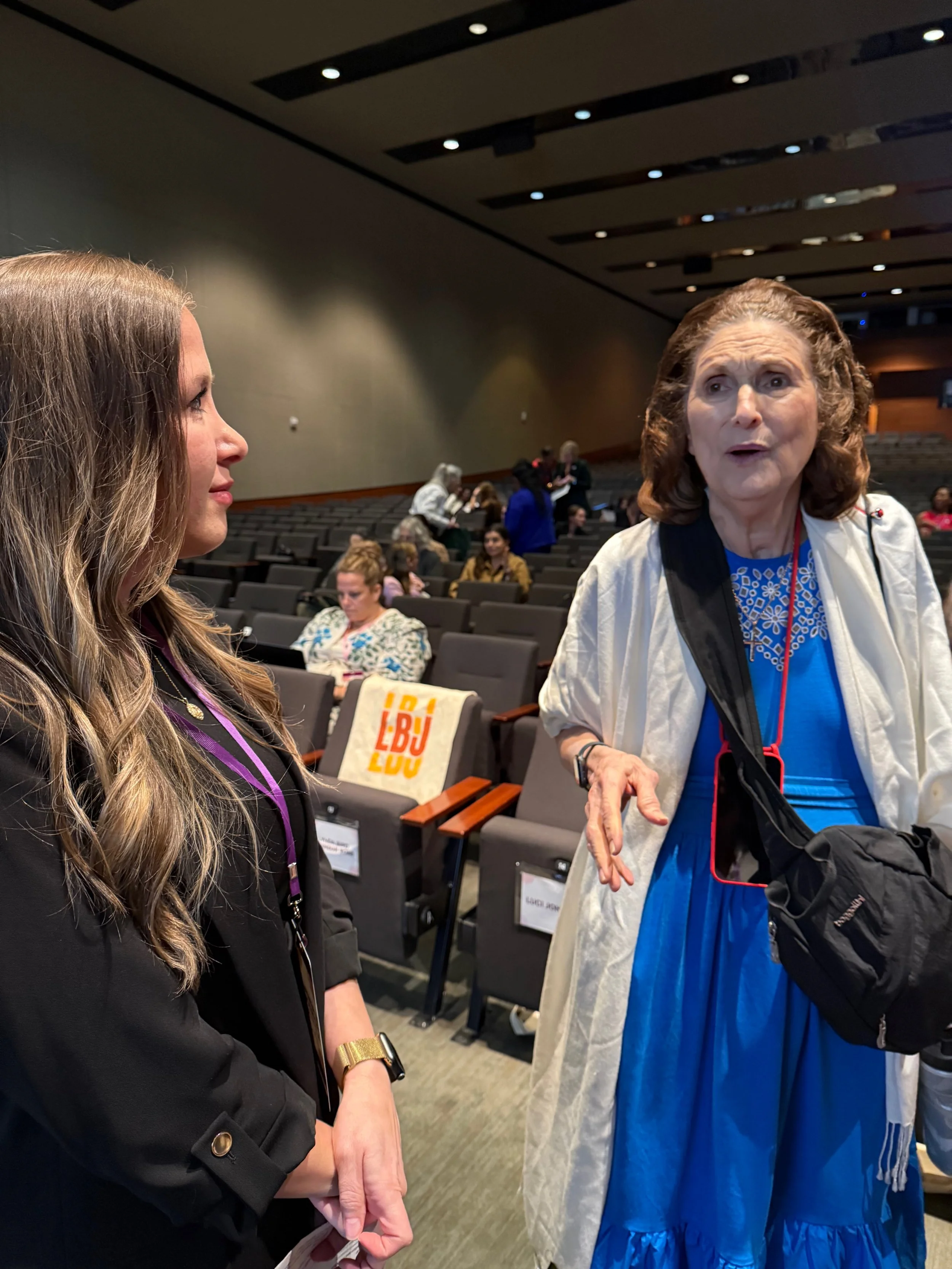 Two women are engaged in conversation in an auditorium, with several other people seated in the background. The woman on the right has gray hair, wearing a white coat and a blue dress, and is gesturing with her hand. The woman on the left has long br