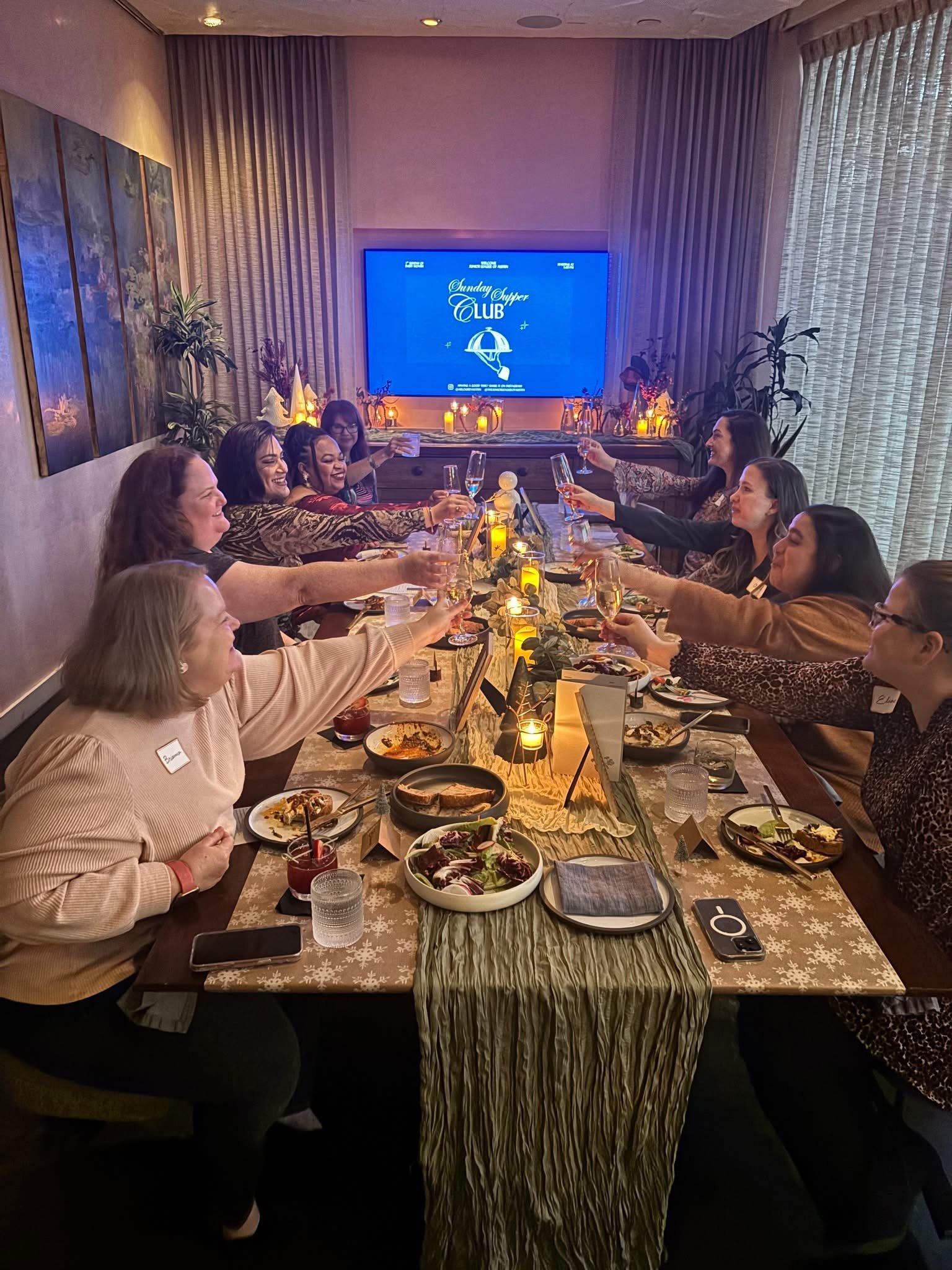 Women gathered around a decorated table for a celebration, toasting with champagne glasses, in a cozy room with candles, plants, and a large TV screen displaying 'Sunday Supper Club'.