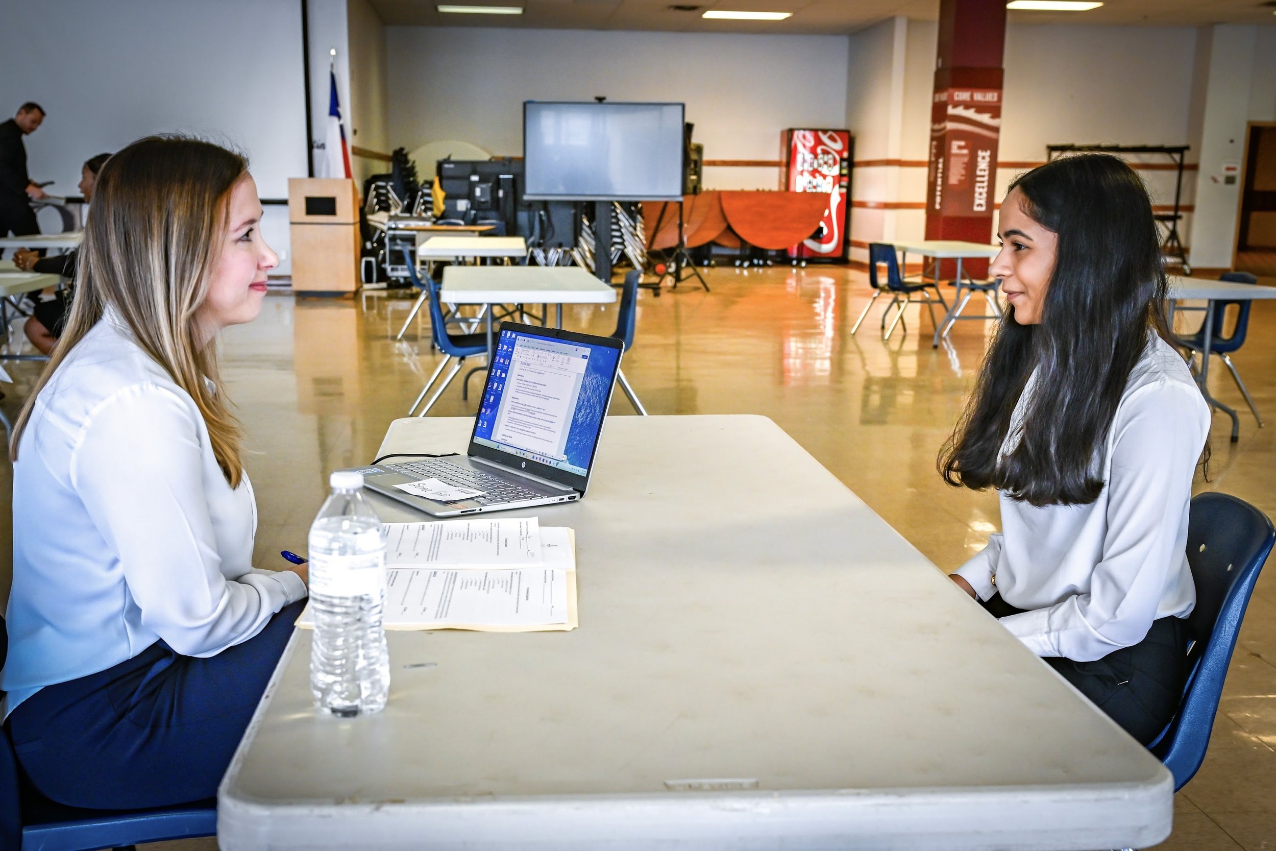 Two women sitting across from each other at a table in a large room, engaged in conversation. One woman has light brown hair and the other has long dark hair. A laptop, water bottle, and papers are on the table.