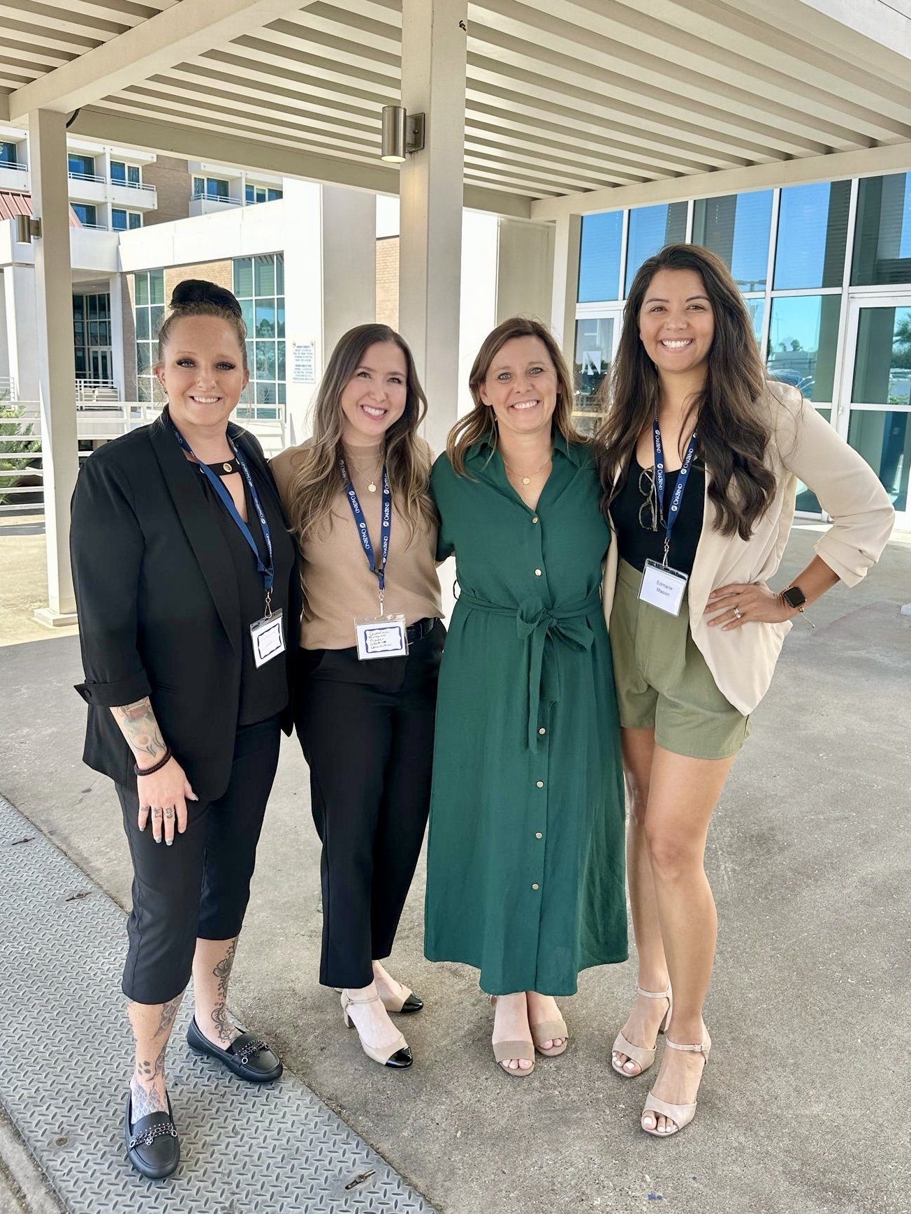 Four women standing together outdoors, smiling, in front of a modern building with large glass windows. They are wearing business casual attire and have name badges. The woman on the far right is wearing a beige blazer and green shorts, the second wo