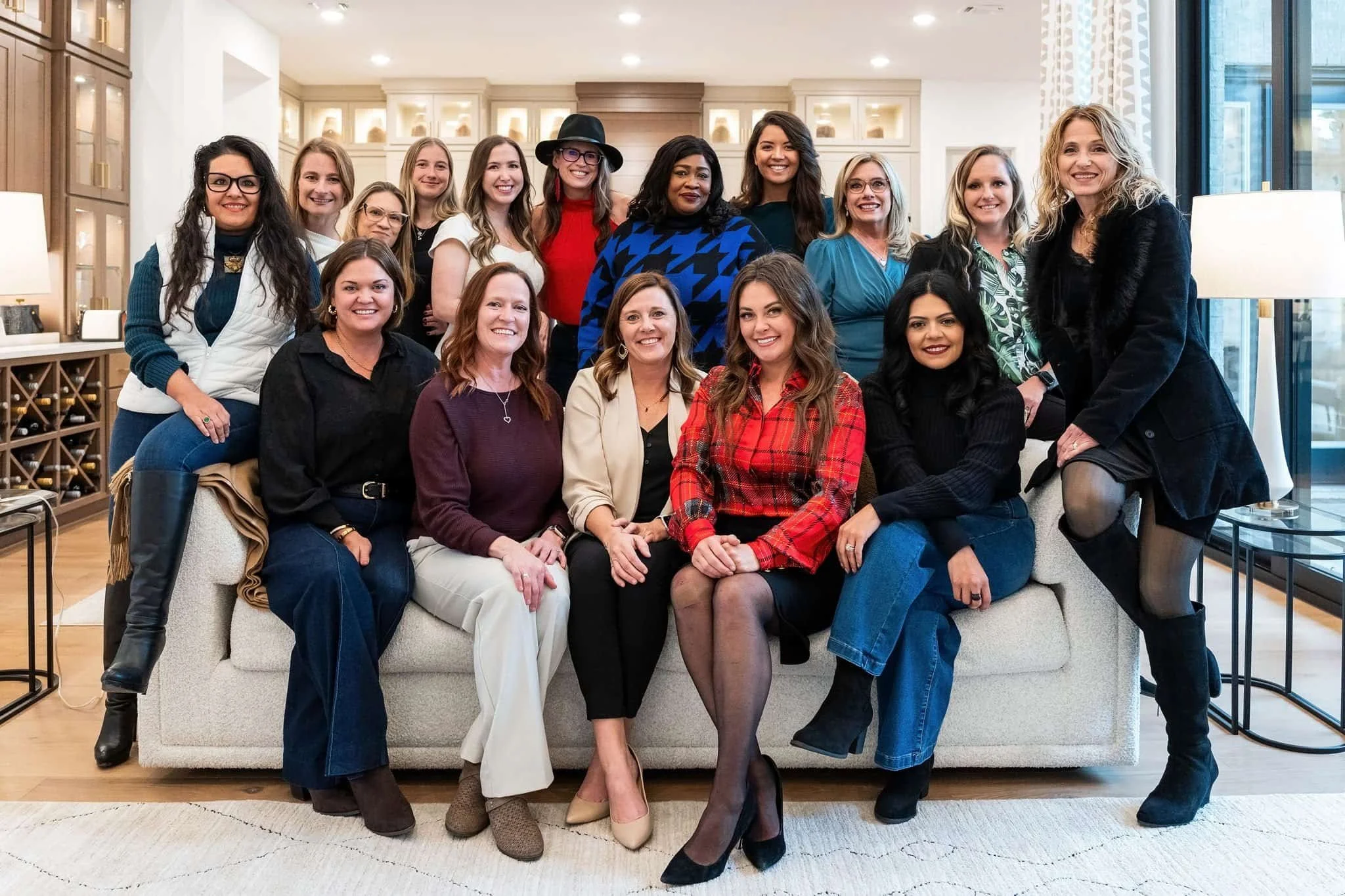 Group of 16 women sitting and standing on a beige sofa in a bright, modern living room with wooden shelves, lamps, and large windows.