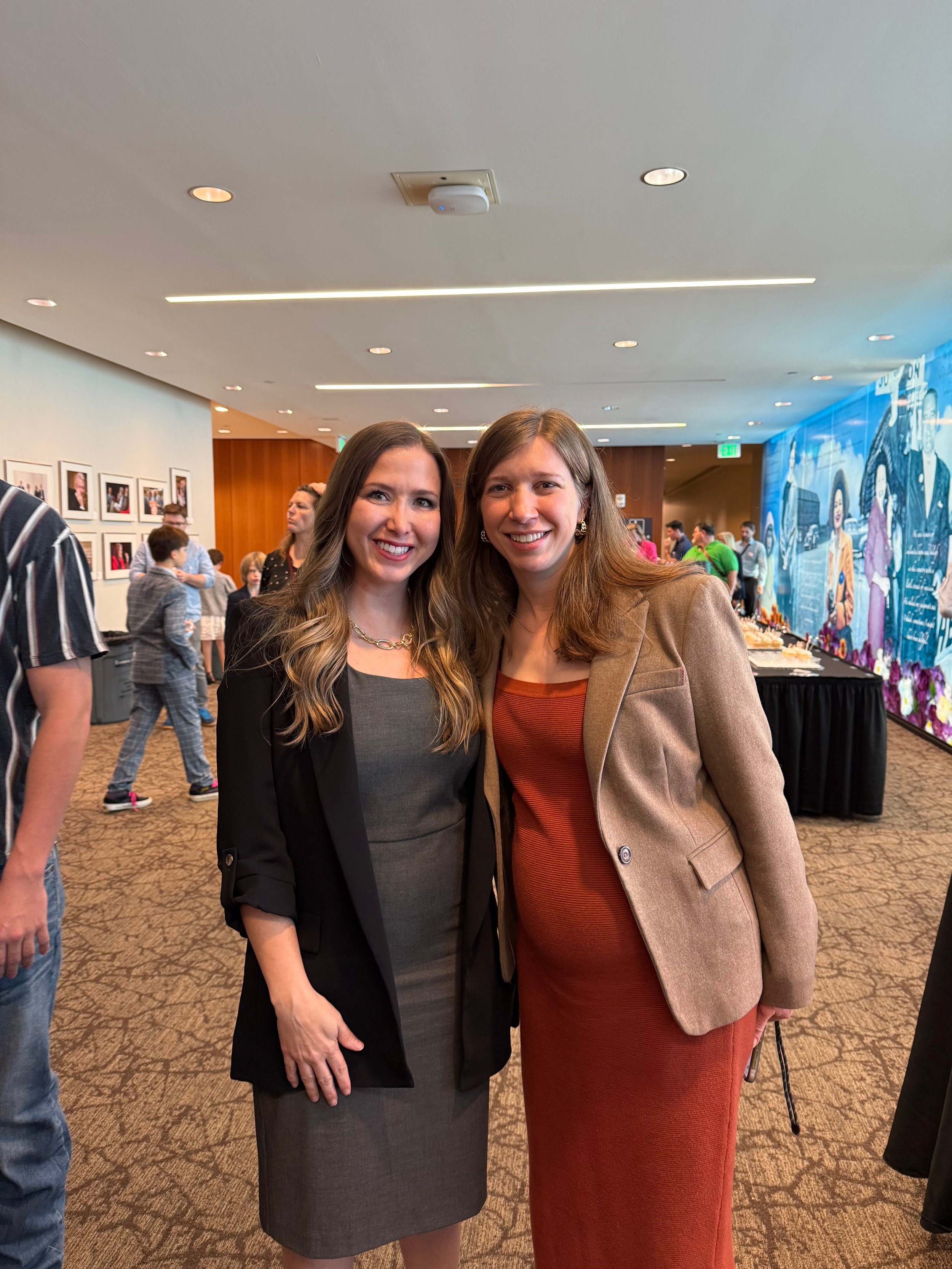 Two women standing together in a conference or event room, smiling at the camera. One wearing a black blazer and gray dress, the other in a beige blazer and red dress. Background includes other people, framed photos, and a blue mural.