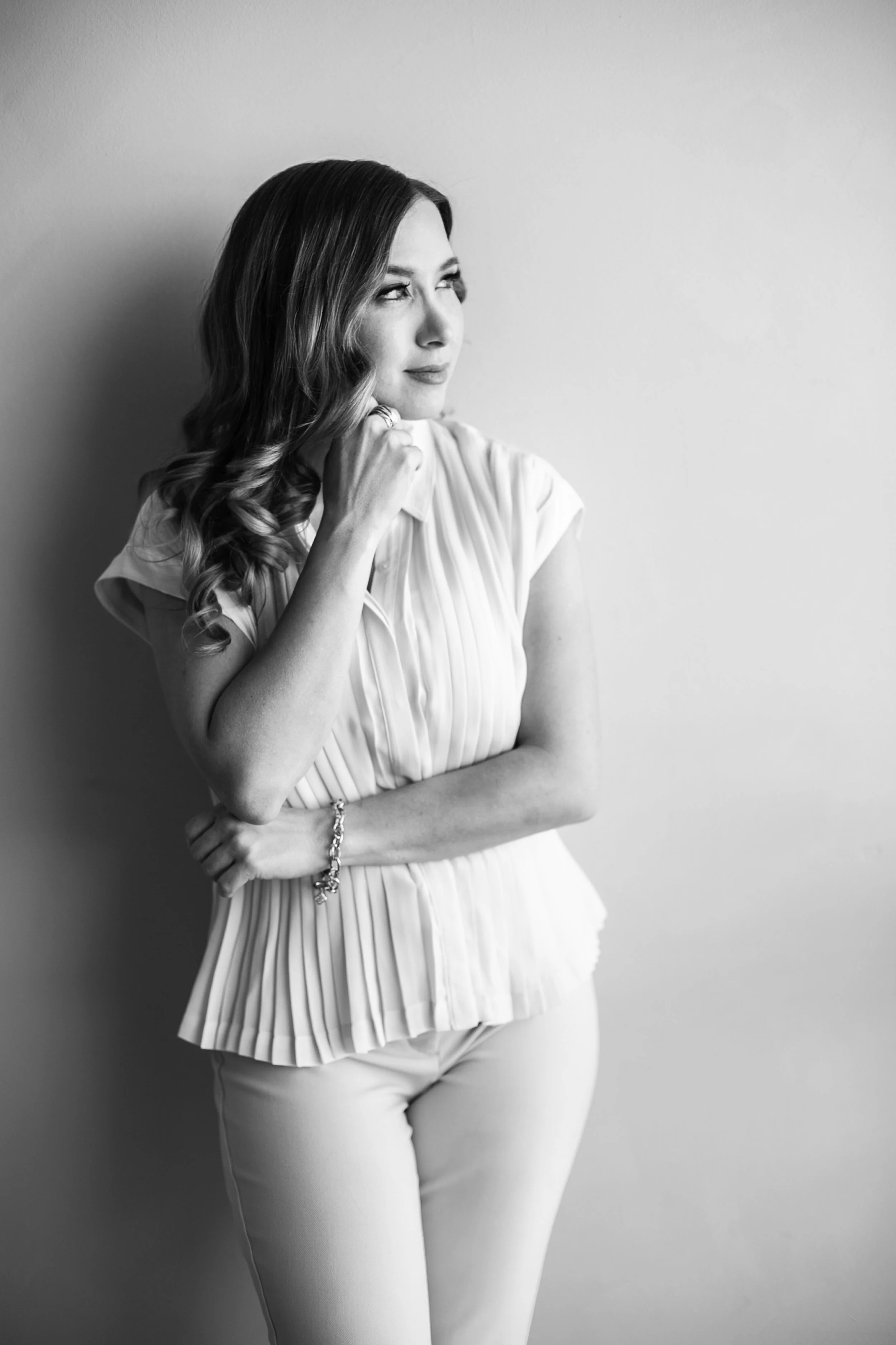 April Guerra with wavy hair, wearing a pleated top and pants, leaning against a wall and looking thoughtfully to her right.