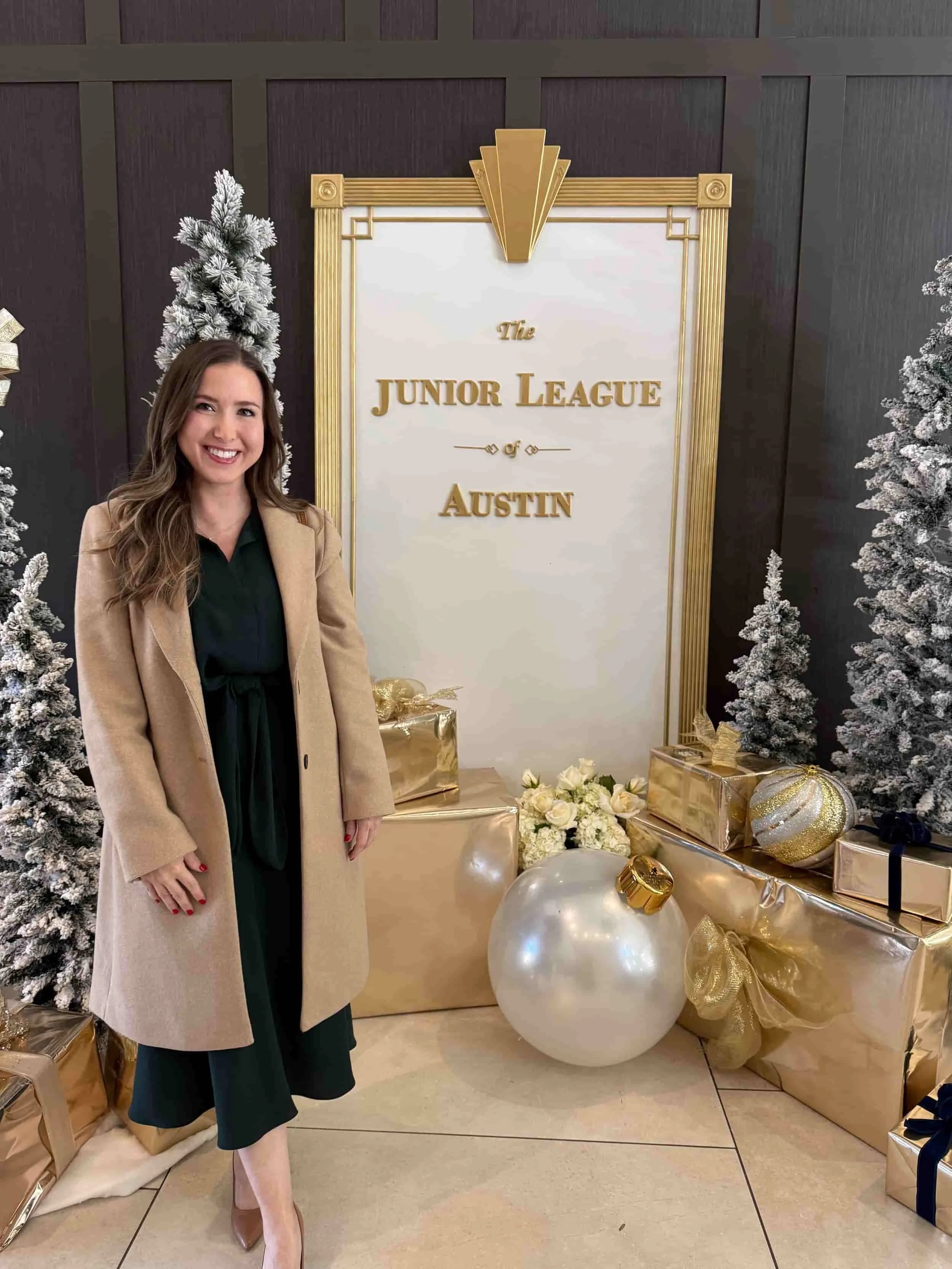 A woman standing in front of a festive winter holiday display with white snow-covered Christmas trees, gold and white ornaments, gold wrapped presents, and a sign that reads 'The Junior League of Austin.'