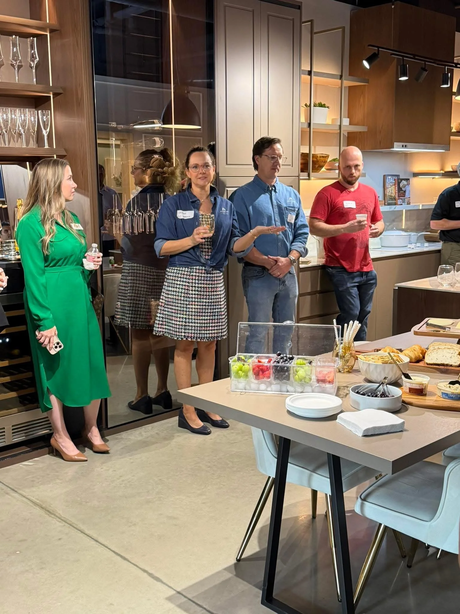People standing in a kitchen area during a social gathering, with a table of snacks and drinks in the foreground.