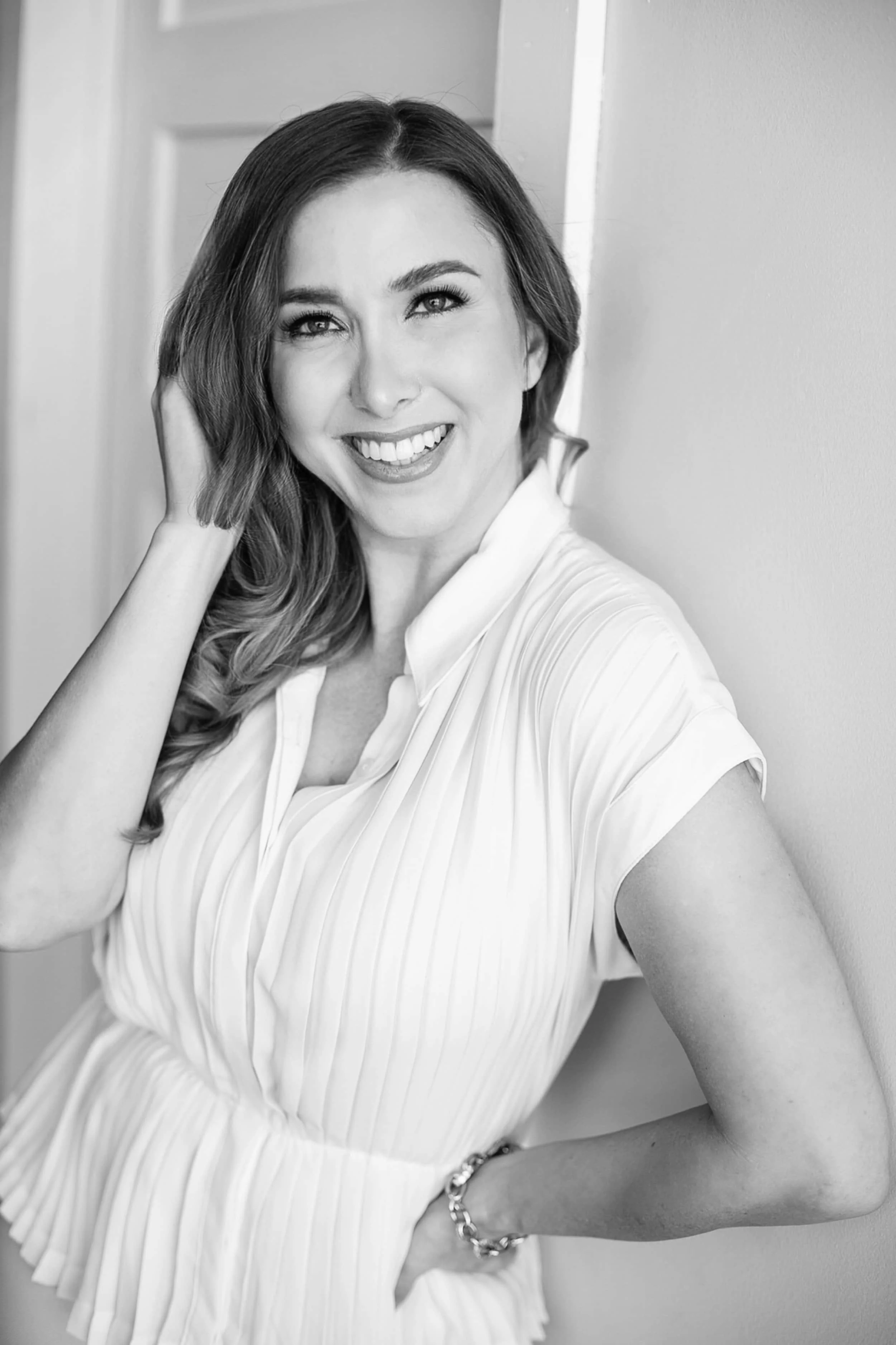 Black and white portrait of a woman, April Guerra, smiling, with one hand touching her hair and the other hand on her hip, wearing a light-colored pleated top, standing indoors near a wall.