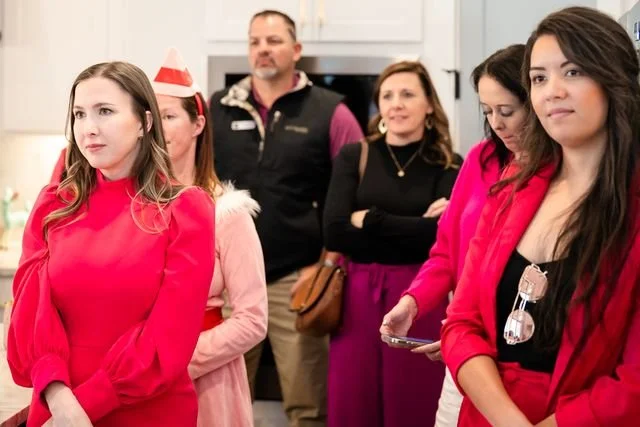 Group of six people standing indoors, some looking serious or thoughtful, one woman wearing festive red attire, others in casual clothing.