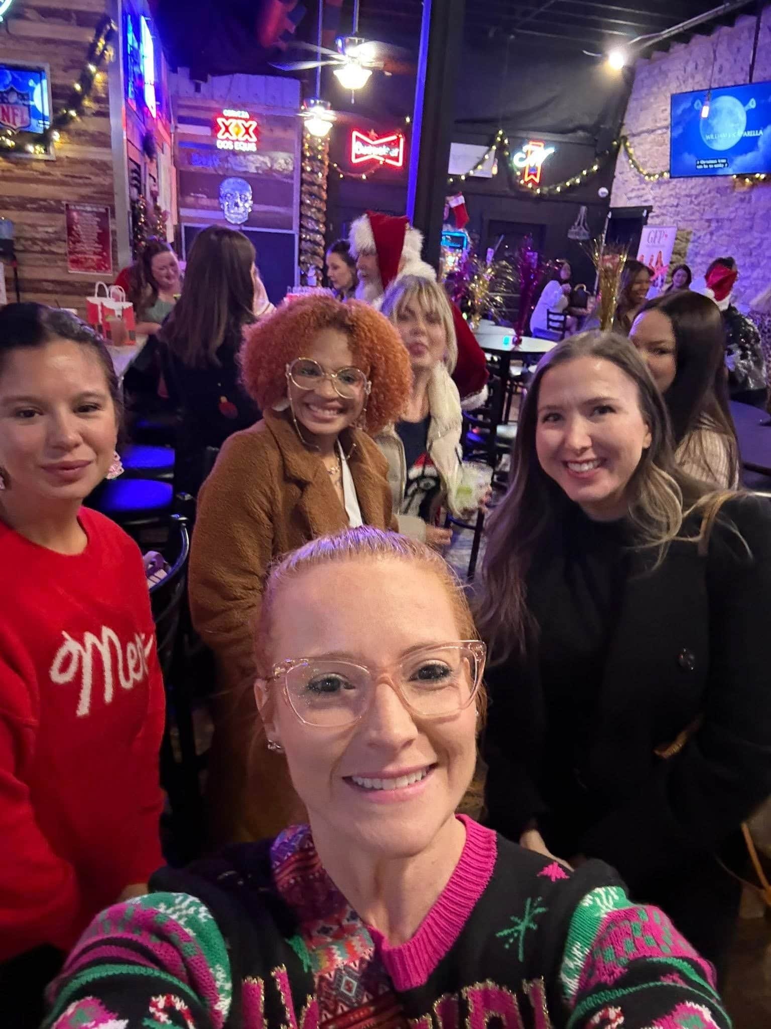 Group of women celebrating Christmas in a festive restaurant, with some wearing holiday sweaters and Santa hats, decorated with Christmas lights and ornaments.