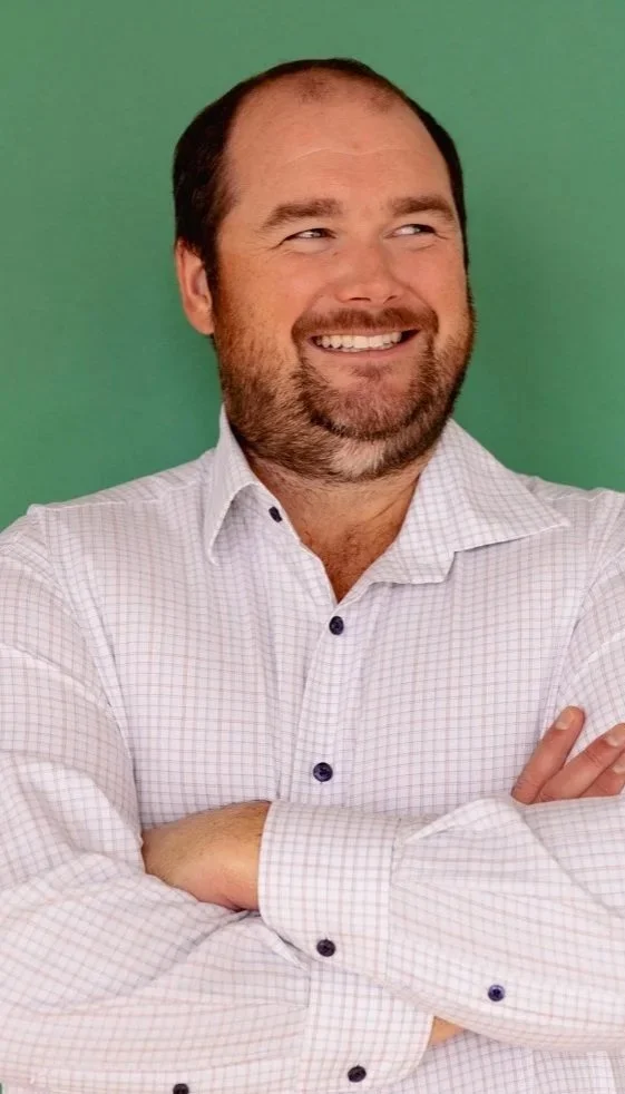 A man with a beard and short hair smiling and crossing his arms, wearing a white checkered shirt, against a green background.