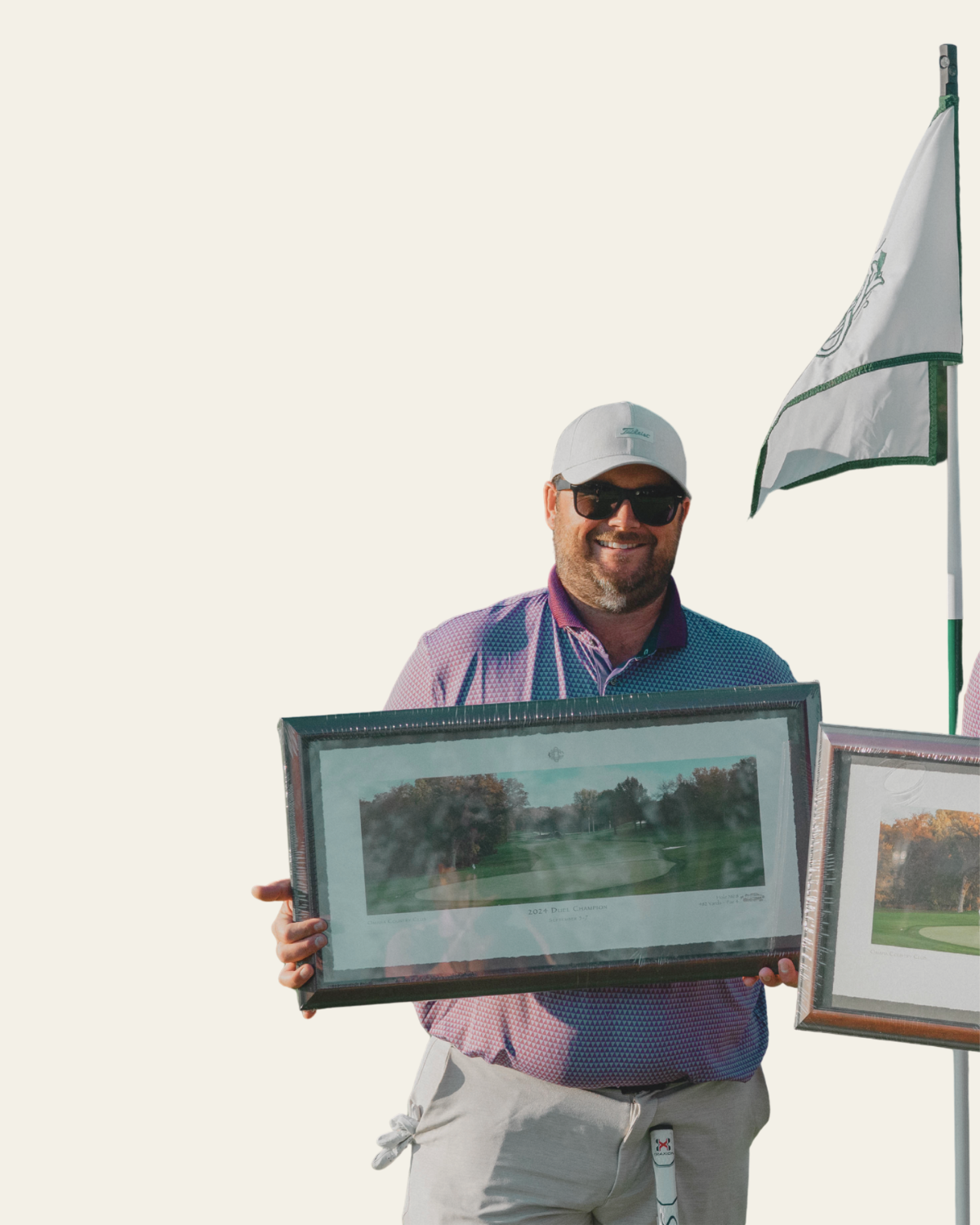 A man smiling, wearing sunglasses, a white cap, and a colorful golf shirt, holding a framed image of a golf course. There is a golf flag in the background.
