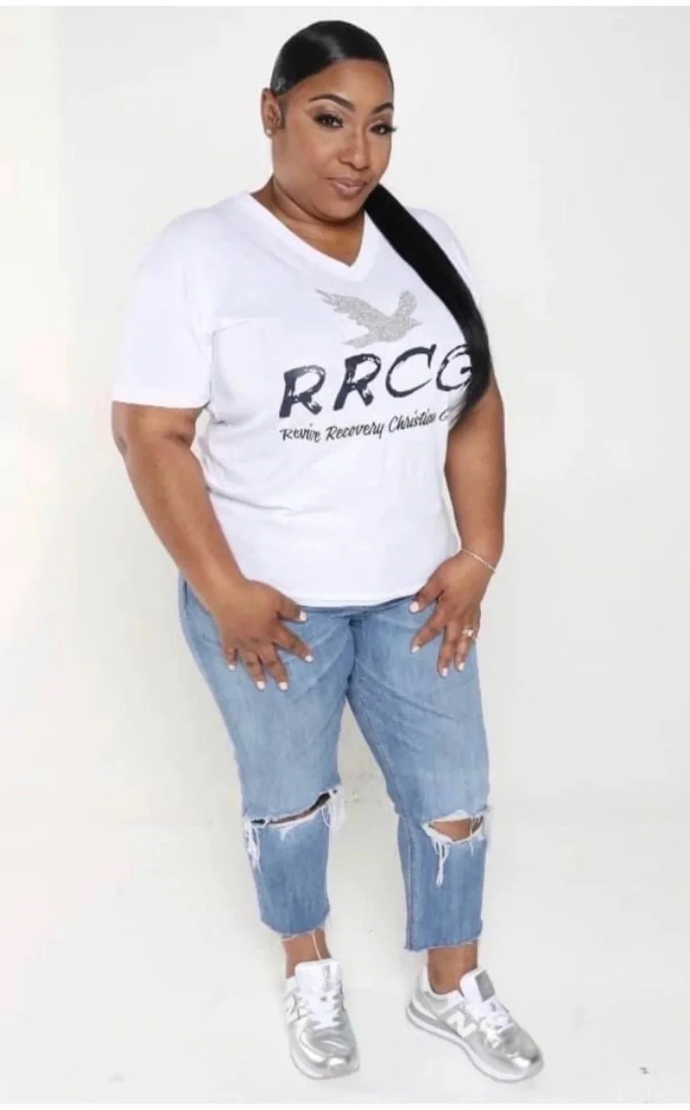 A woman standing against a plain white background wearing a white t-shirt with the text 'RRCC' and a dove graphic, ripped jeans, and silver sneakers.