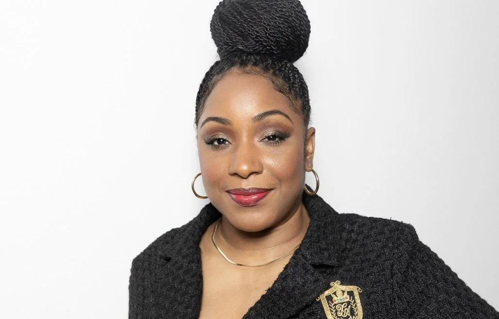 Portrait of a confident African American woman with braided hair styled in a high bun, wearing gold hoop earrings, a black textured blazer with a crest pin, and red lipstick, against a plain white background.