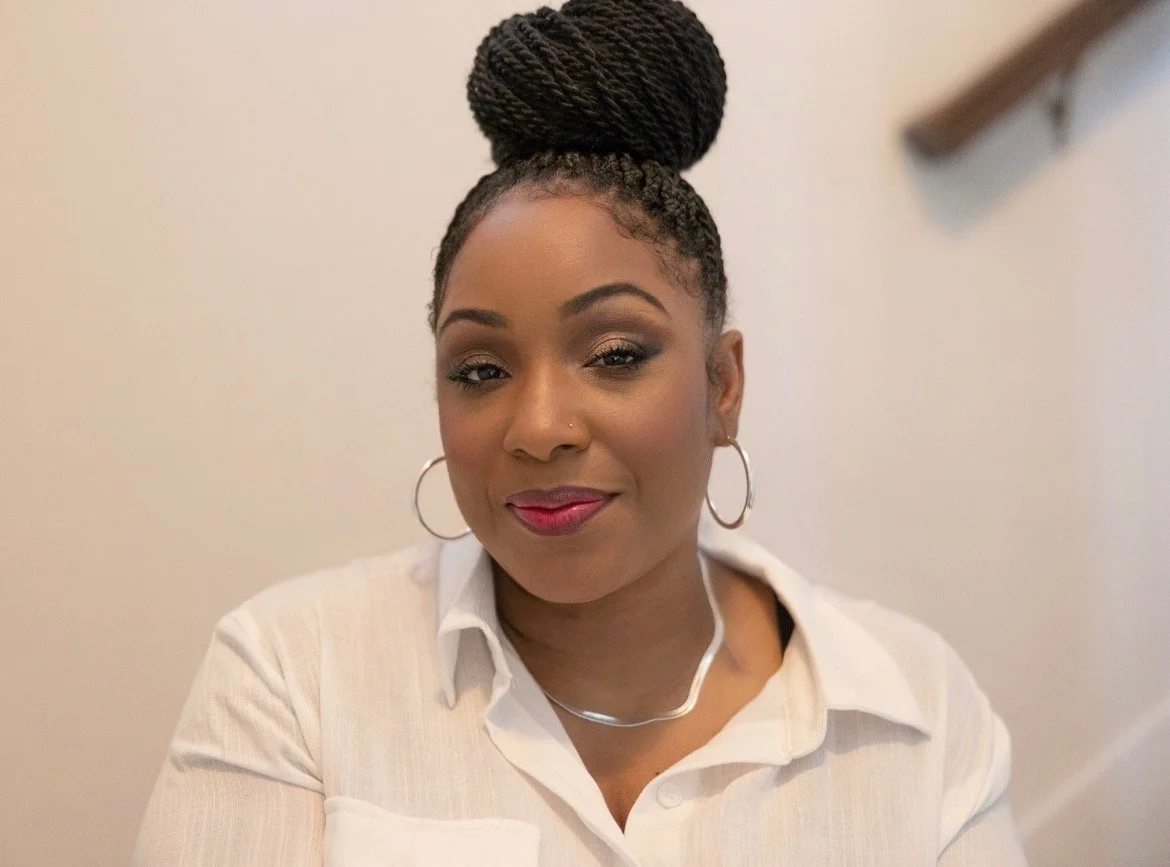A woman smiling with her hair styled in a high braided bun, wearing hoop earrings, a white shirt, and red lipstick, against a plain wall background.