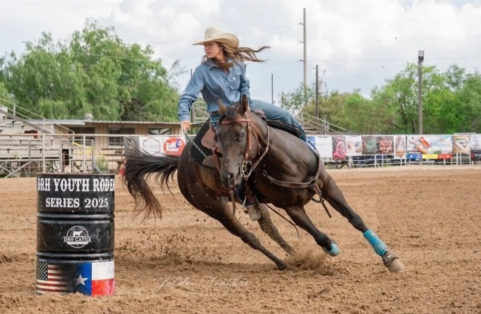 A young girl wearing a cowboy hat and denim shirt rides a horse around a barrel during a youth rodeo event. The barrel is labeled with "Future Youth Rodeo Series 2025" and has a small Texas flag painted on it. The arena has a dirt floor, with trees, banners, and picnic tables in the background.