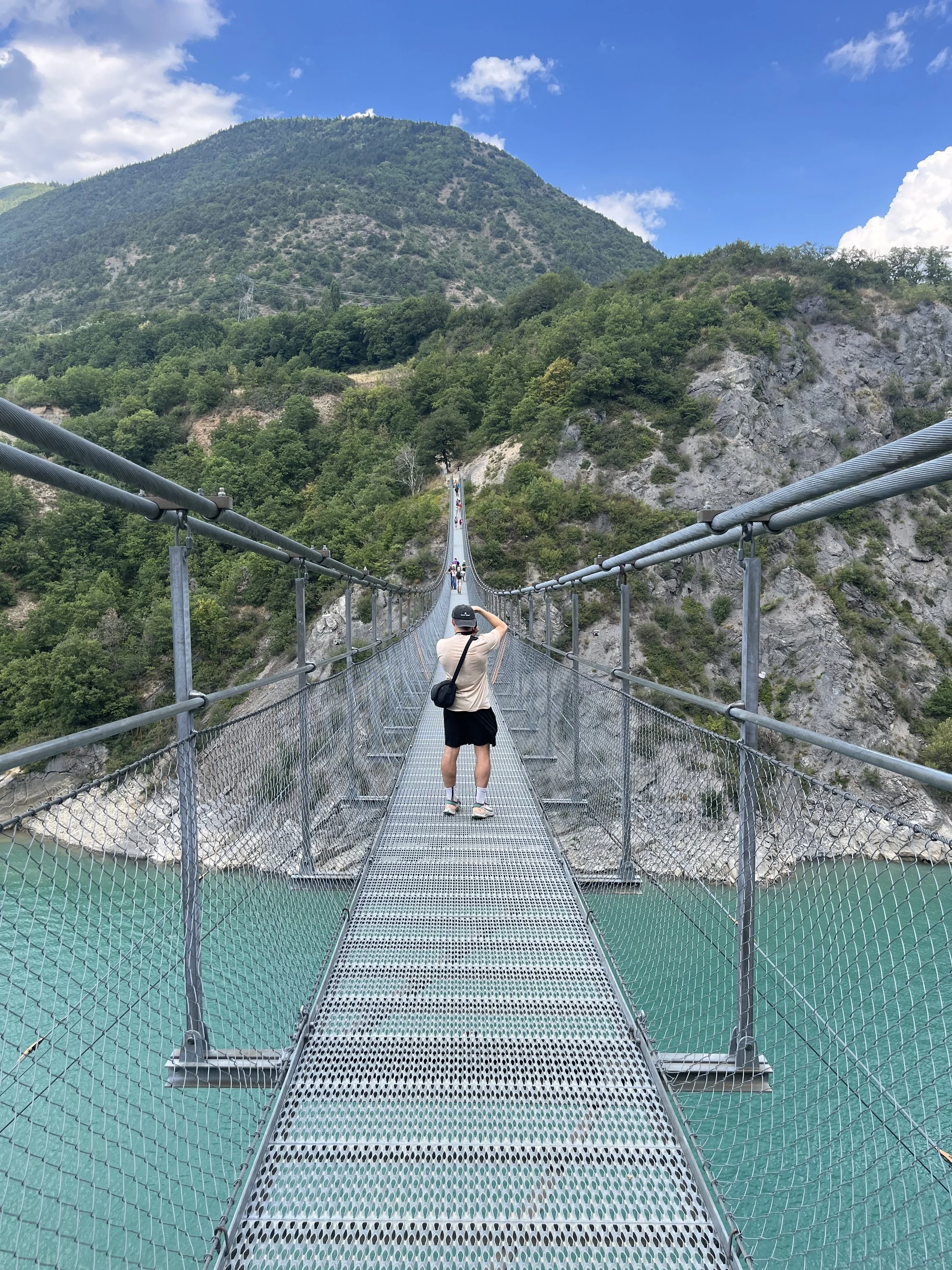 Personne photographiant un pont suspendu métallique au-dessus d'une rivière turquoise, avec une montagne verte en arrière-plan.