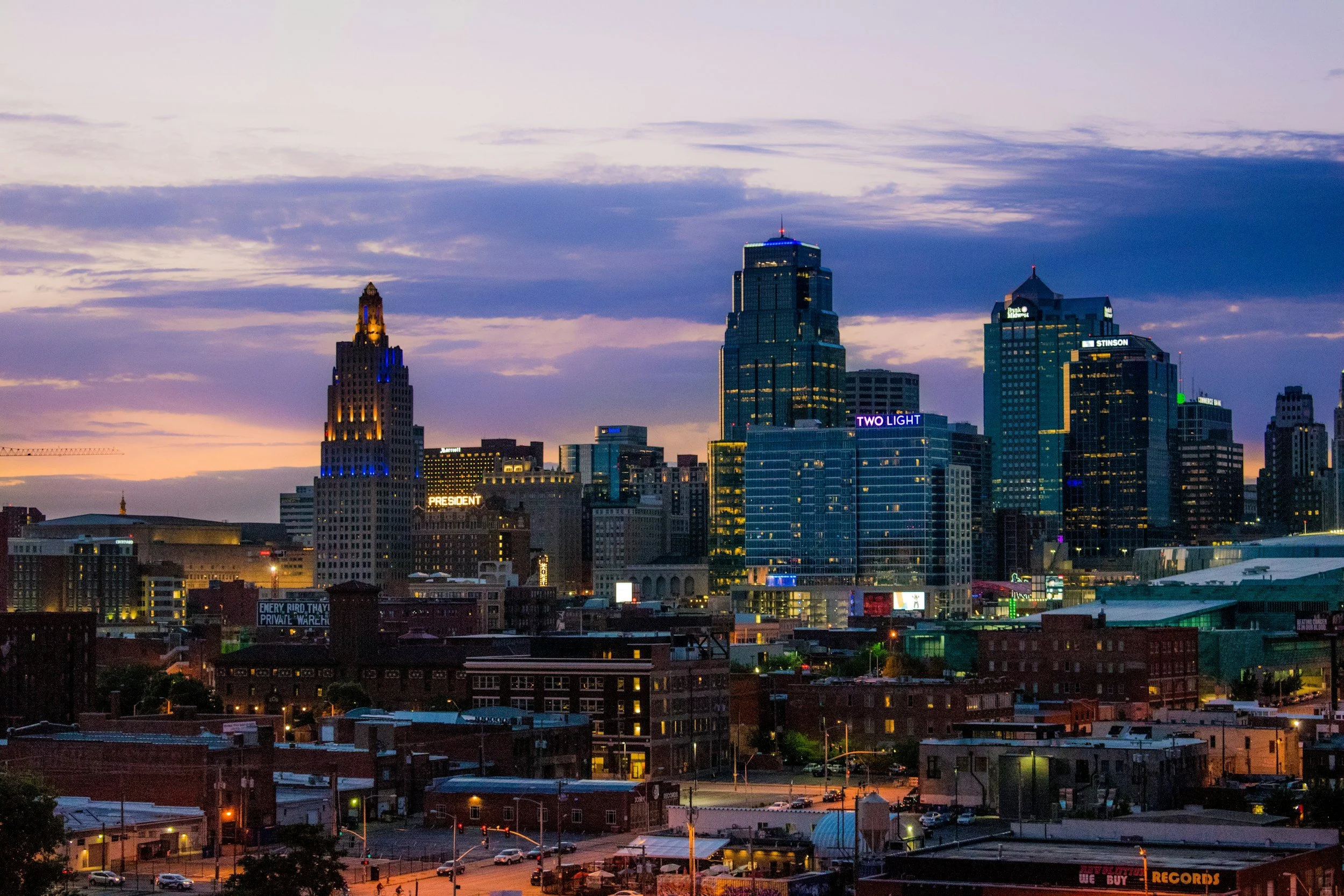 Nighttime city skyline with tall buildings, some illuminated signage, and colorful clouds in the sky.