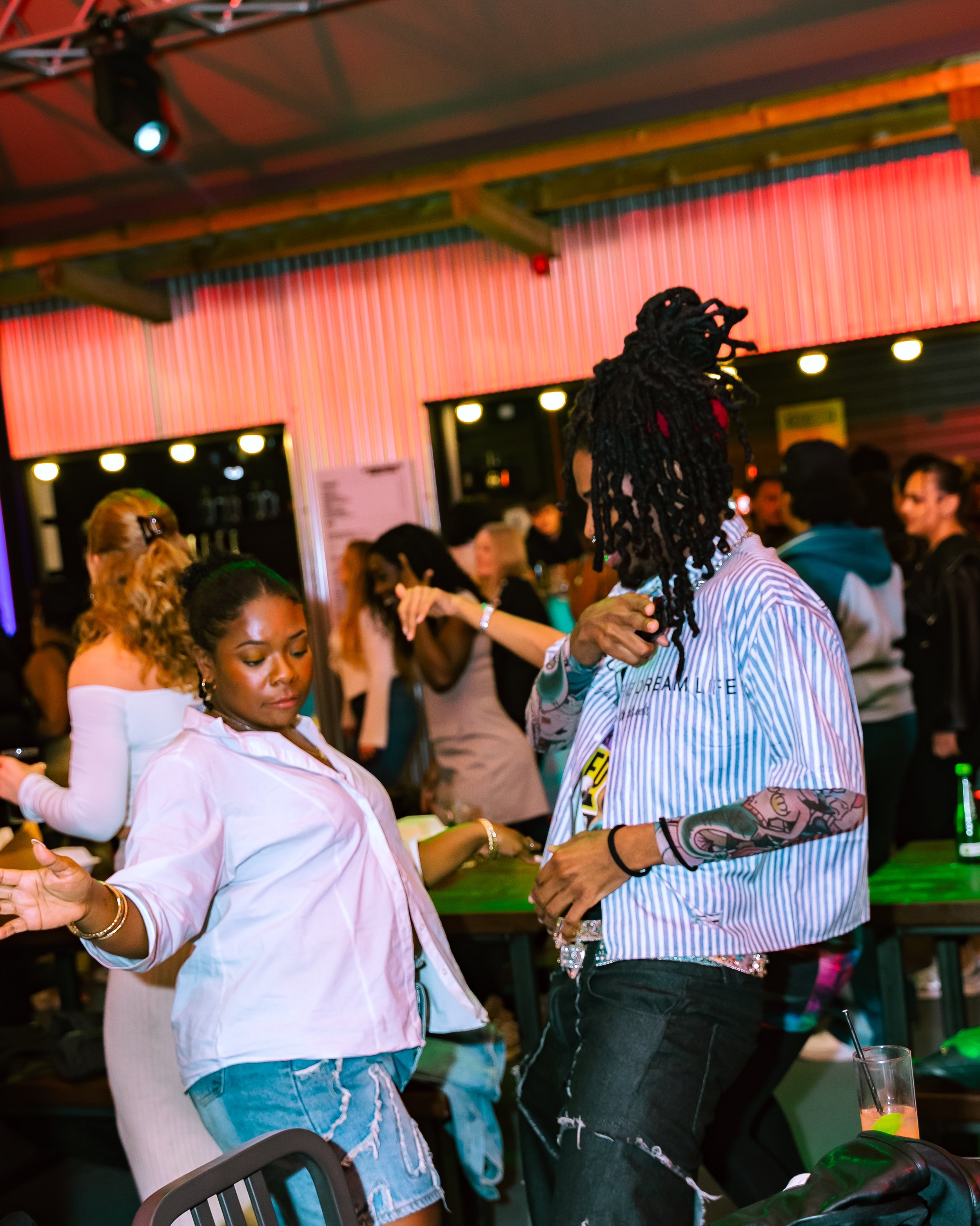 People dancing and socializing at a nightclub or party venue with colorful lighting and a bar in the background.