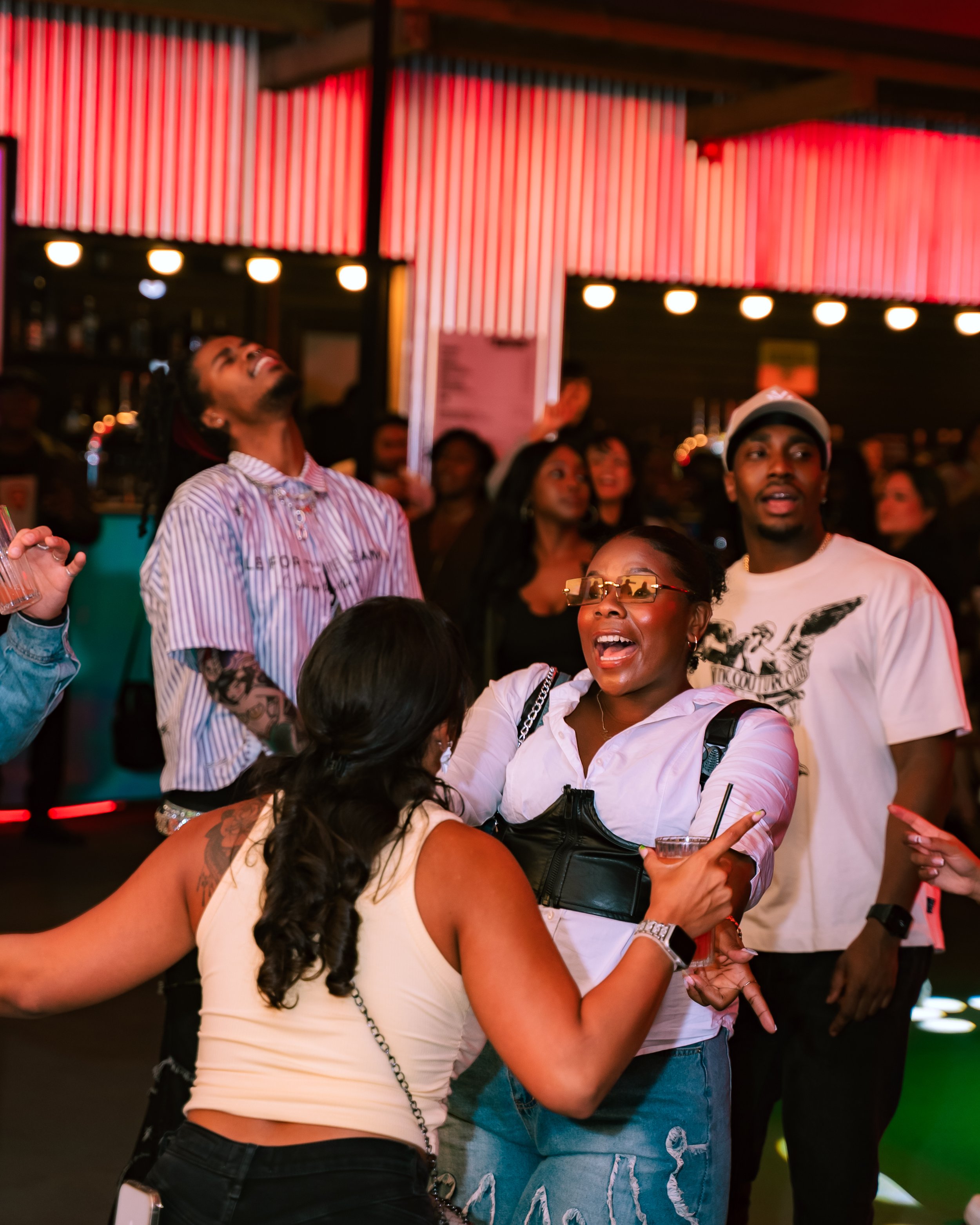 People at a lively social event, some engaged in animated conversations, others looking up or around, all under red neon lighting.