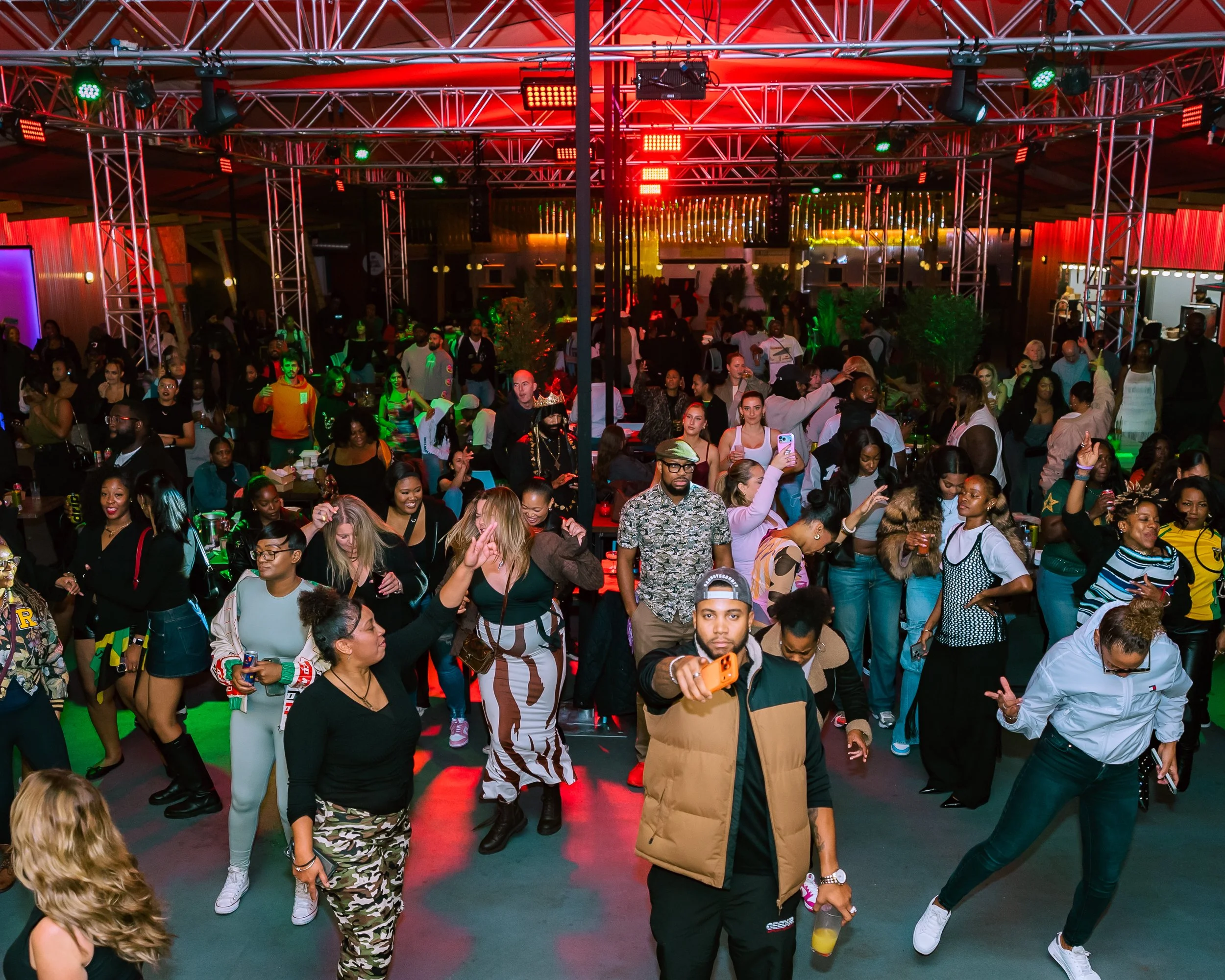 Crowd of people dancing and socializing under colorful stage lighting at an indoor party or nightclub.