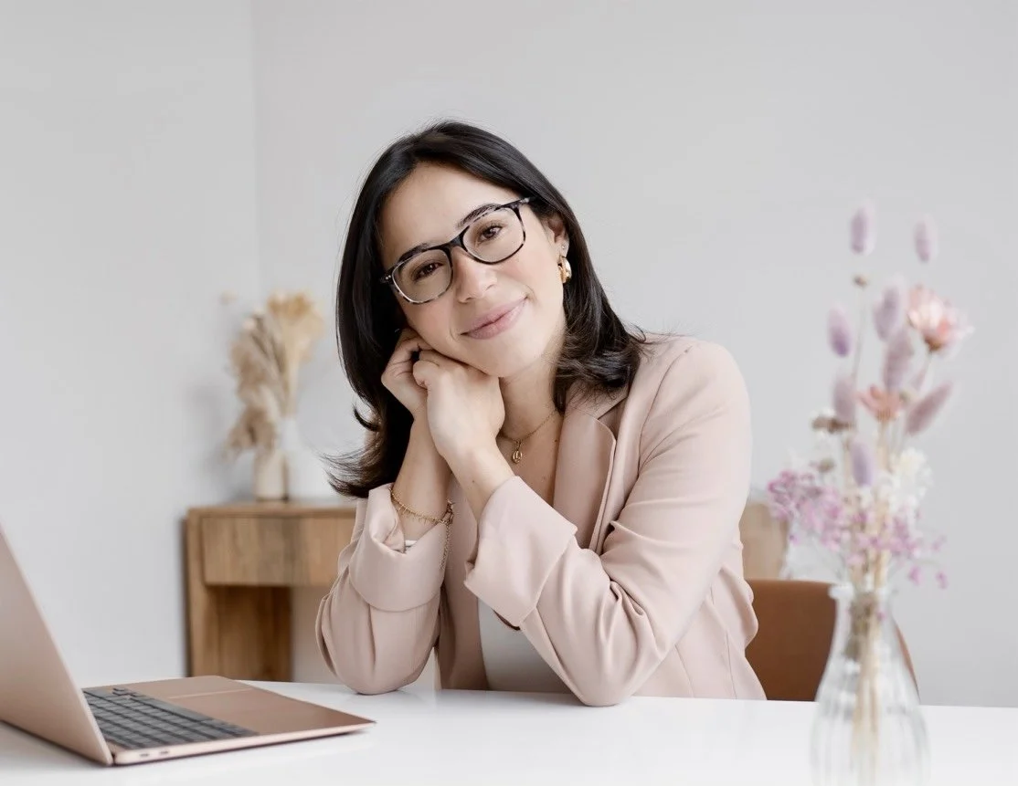 A woman with dark hair, glasses, and gold jewelry sitting at a desk with a laptop and a vase of flowers, smiling softly at the camera.