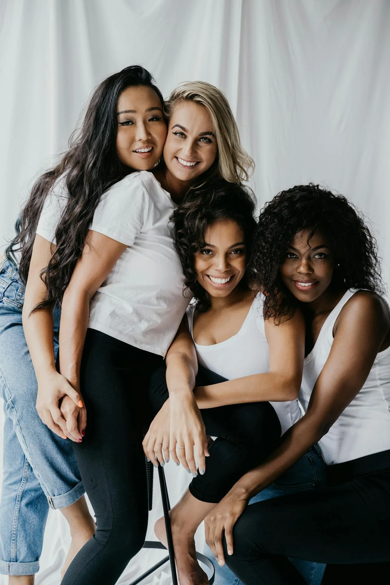 Four women of diverse ethnicities smiling and posing together in a studio with a white backdrop, dressed in casual white tops and dark pants.