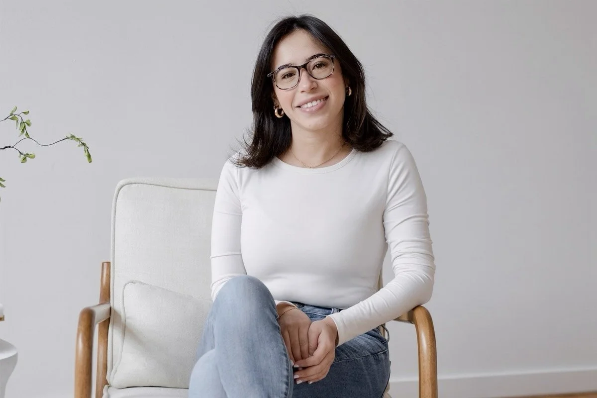 A woman with dark hair and glasses sitting on a cream-colored chair, smiling at the camera, wearing a white long-sleeve shirt and blue jeans, in a minimalistic room with a potted plant to the side.