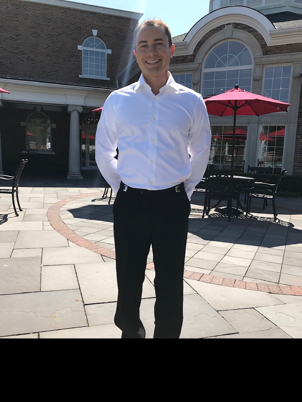 A man in a white dress shirt and black trousers standing outdoors on a patio with tables and red umbrellas.