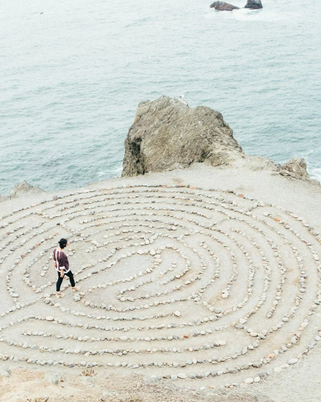 A person walking through a spiral rock garden near the ocean, with a large rock formation in the background.