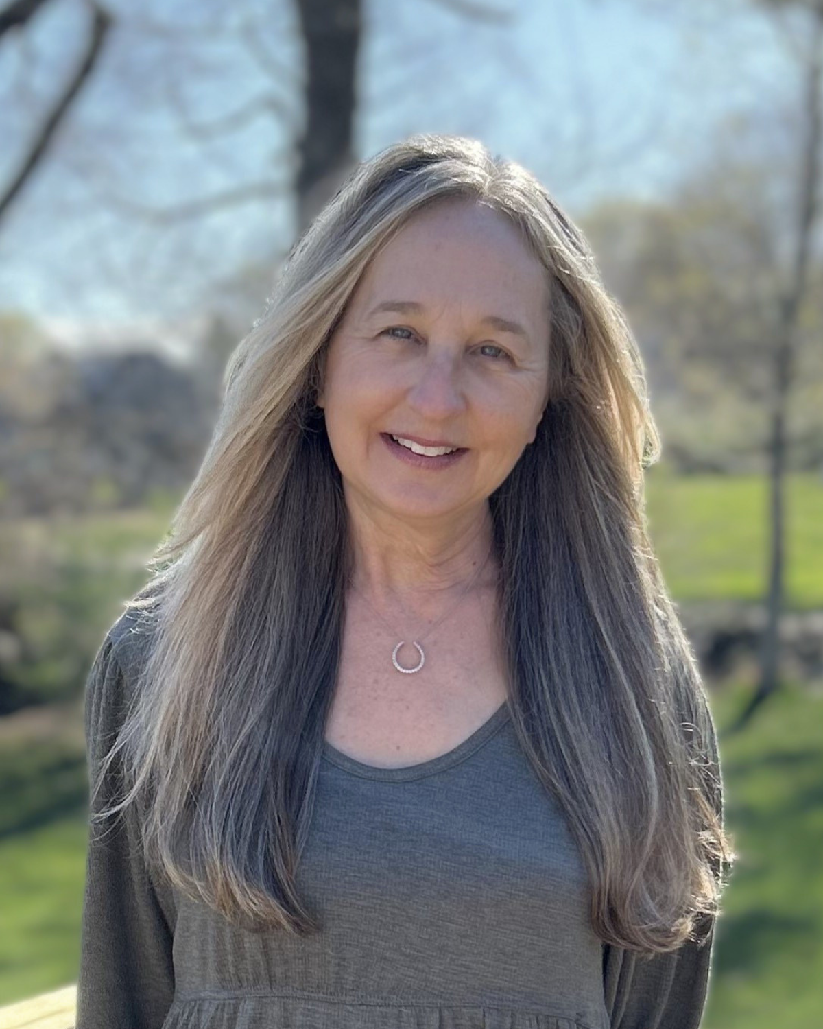 A middle-aged woman with long, gray hair, smiling outdoors on a sunny day, wearing a gray top and a silver circular necklace, with trees and a grassy area in the background.