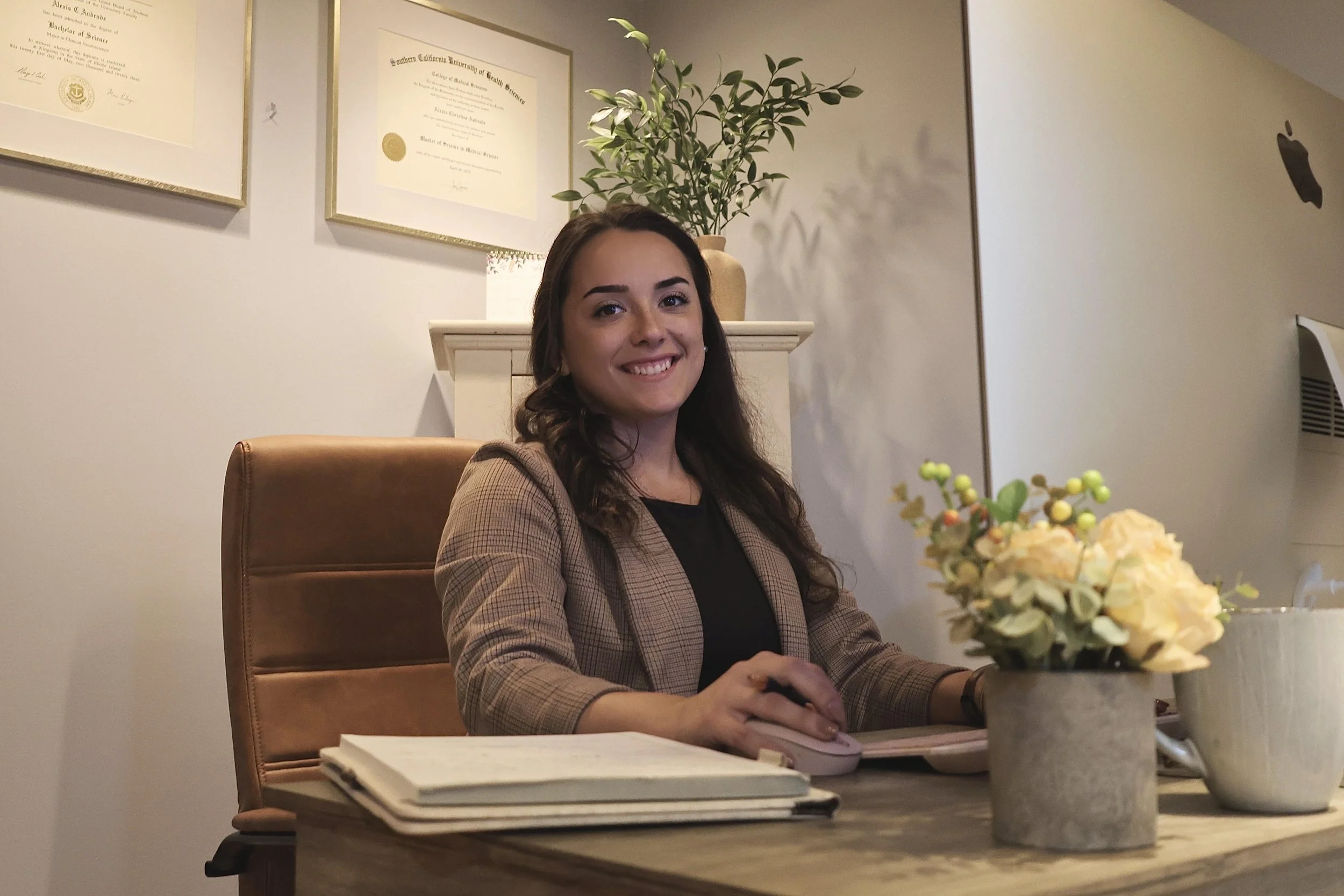 A young woman with long brown hair, smiling, at a desk wearing a black shirt and a blazer in an office setting.  