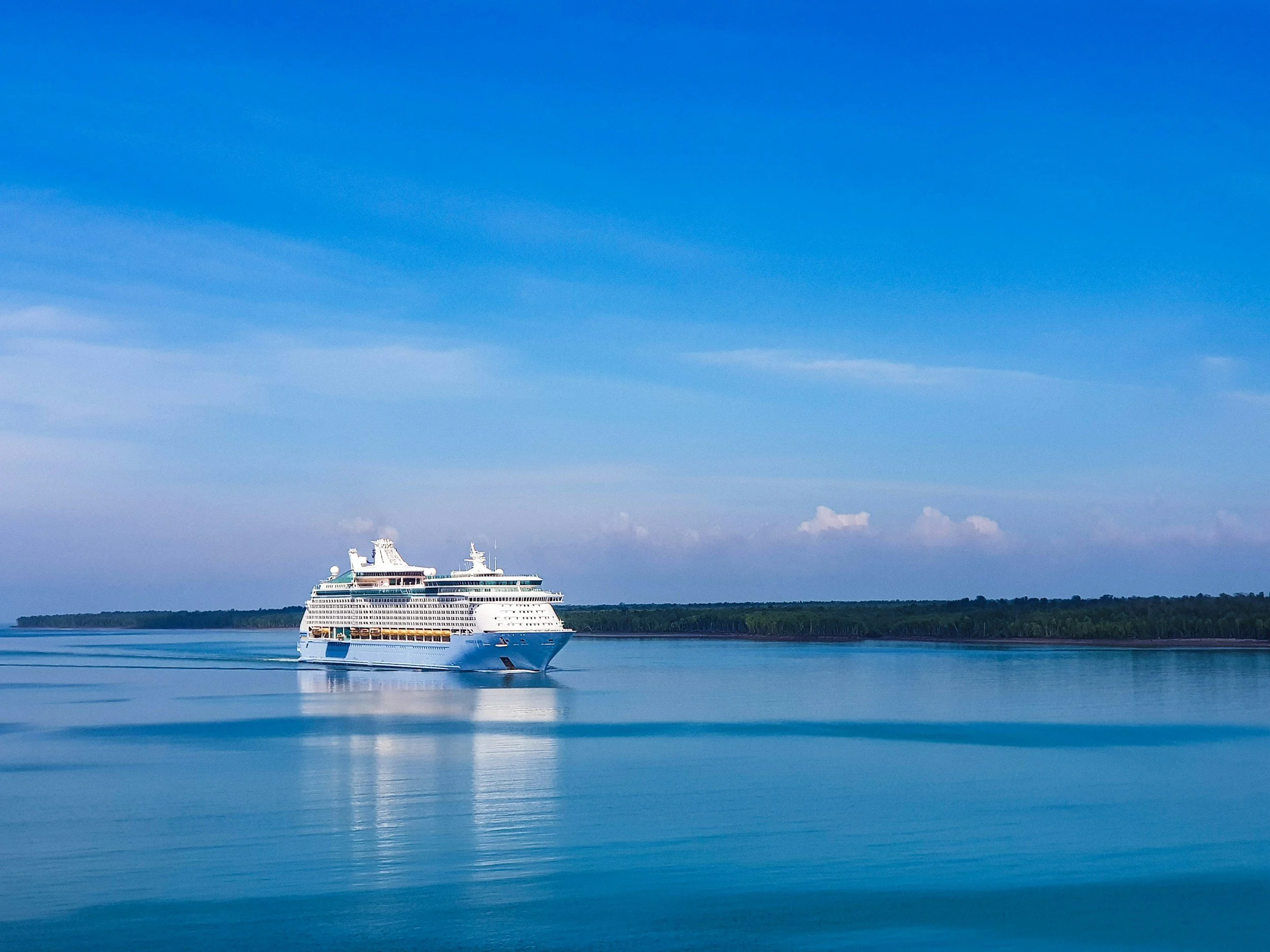 A large white cruise ship sailing on calm blue water against a distant green shoreline under a partly cloudy sky.