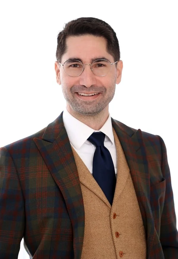 A man with glasses, dark hair, and a beard, smiling, wearing a plaid blazer, tan vest, white shirt, and navy tie, against a white background.
