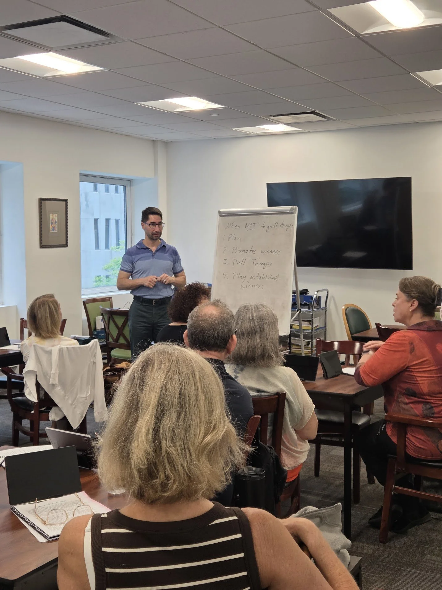 A man giving a presentation to a group of people in a conference room. The presenter is standing by a whiteboard with handwritten notes and a large flat-screen TV on the wall behind him. The room has large windows, wooden tables, and chairs, with some attendees taking notes or listening attentively.