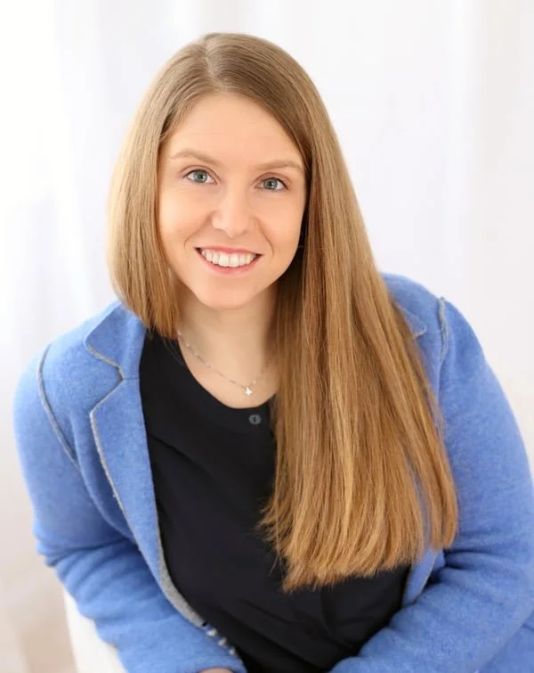 A young woman with long red hair, blue eyes, and a fair complexion, smiling, wearing a black top and a blue jacket, sitting indoors in front of a white background.