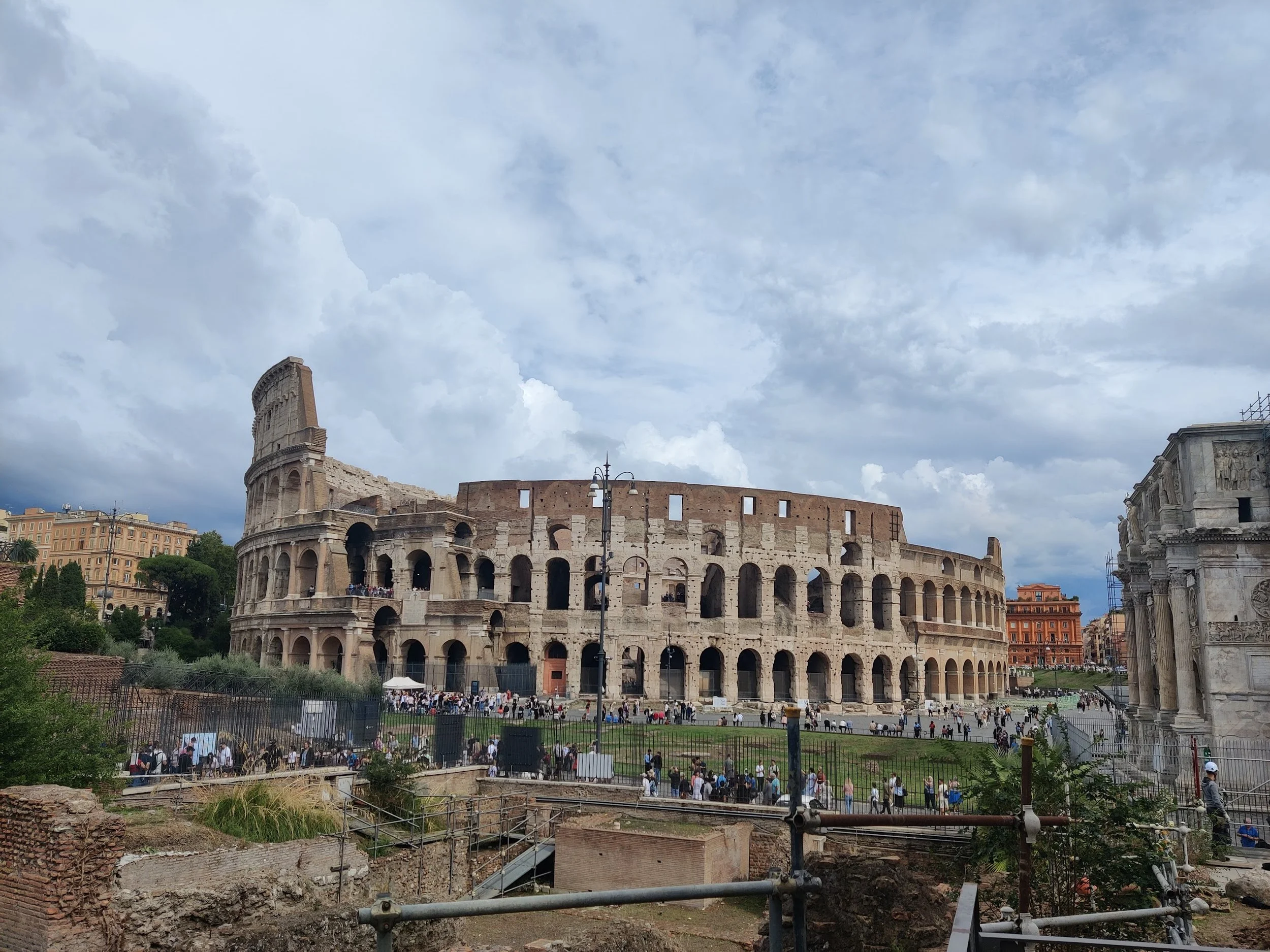 View of the Roman Colosseum with crowds of visitors, surrounded by historic buildings and cloudy sky.