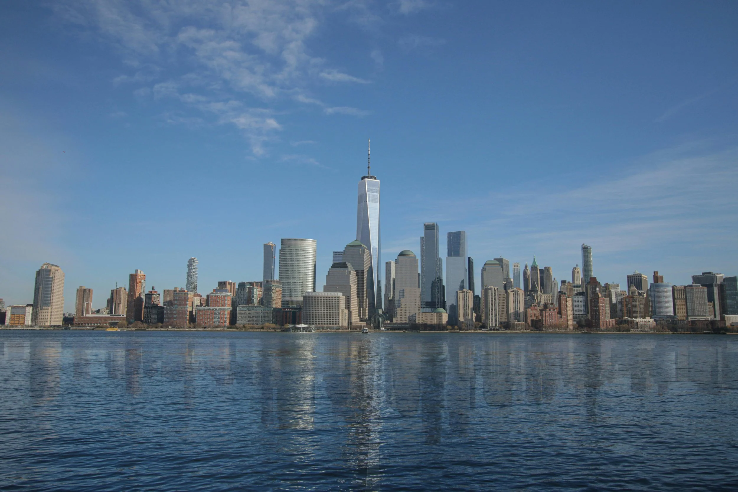 View of New York City skyline with One World Trade Center over water under a partly cloudy sky.