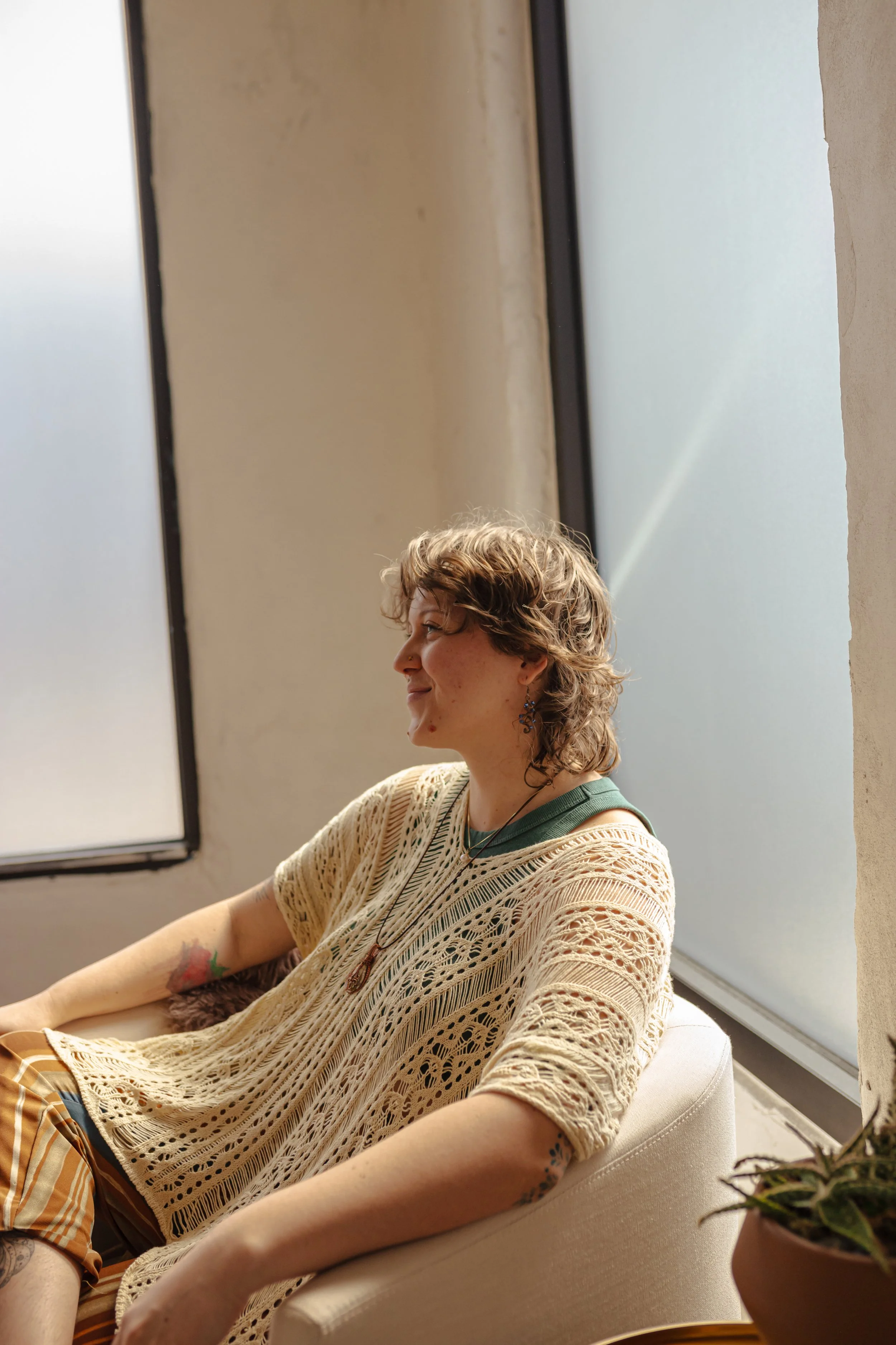 A woman with short, curly hair sitting on a white couch near large frosted windows, wearing a beige crocheted top and patterned shorts, with tattoos on her arms and earrings.