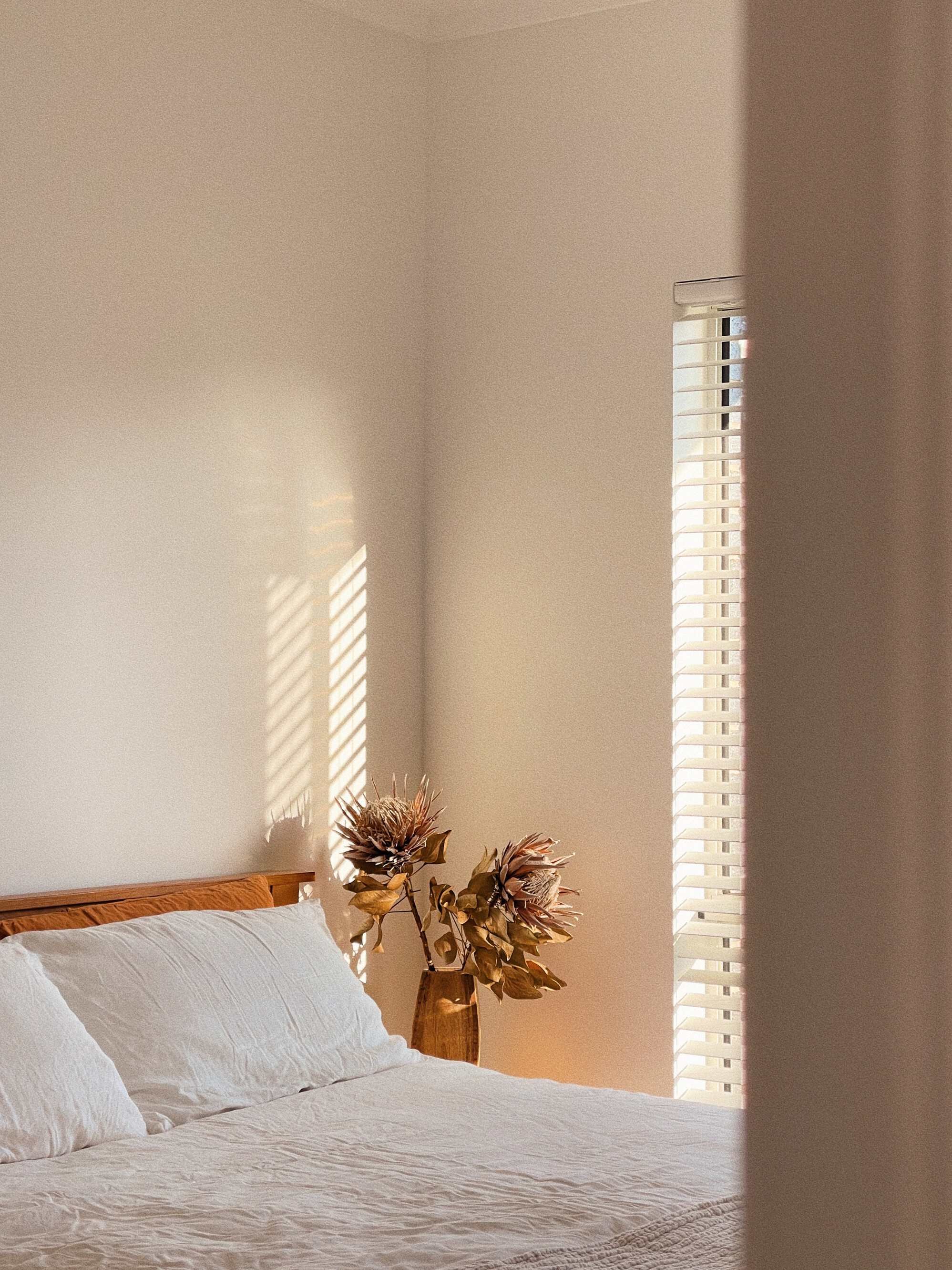 A bedroom with a bed, white bedding, and two pillows. A wooden nightstand with a vase holding dried flowers. Sunlight passing through horizontal blinds creates shadow patterns on the wall.
