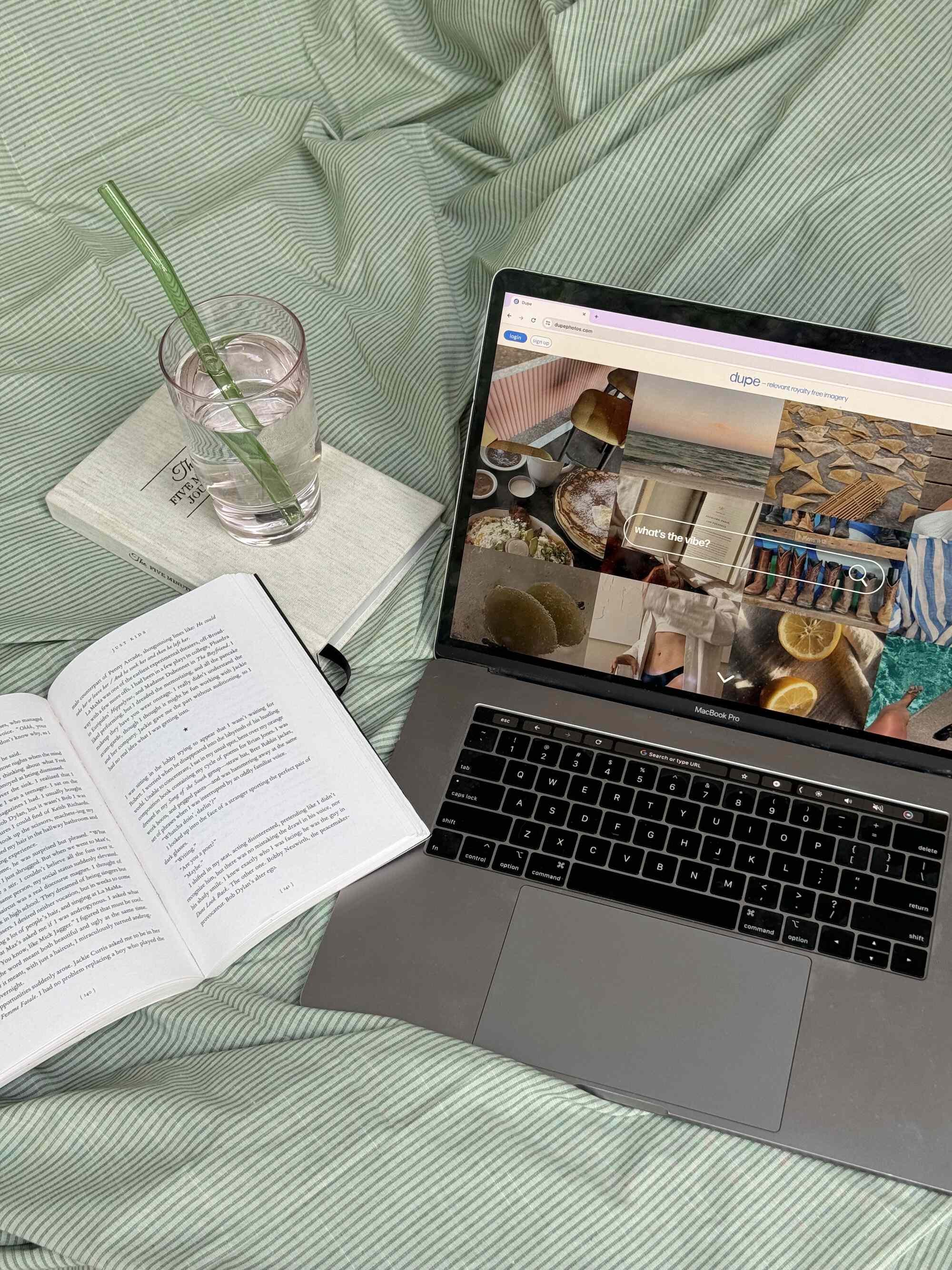 Laptop on a bed with a website open, an open book, a glass of water with a green straw on a book, and a green and white striped bedsheet.