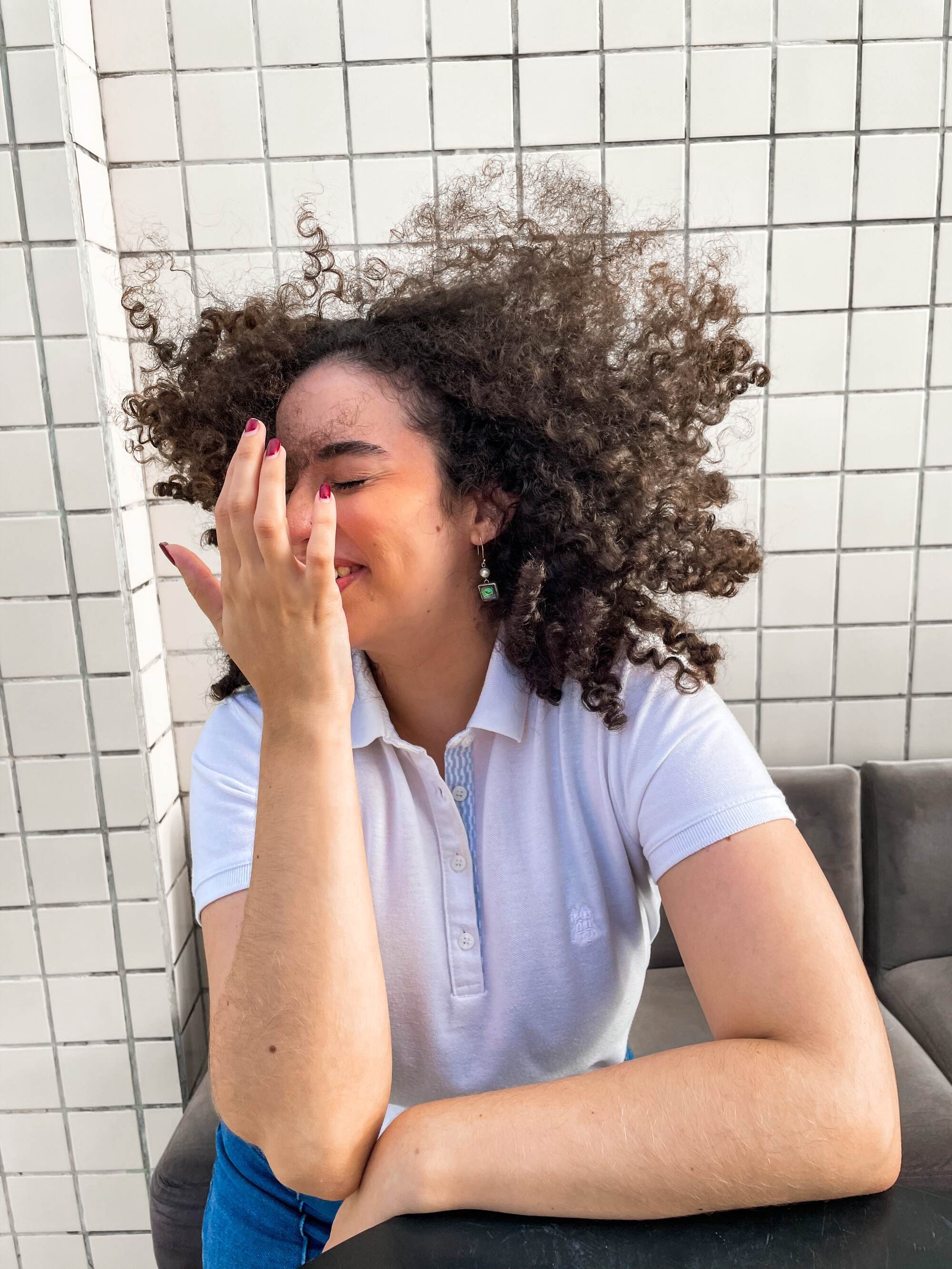 Woman with curly hair sitting at a table, covering part of her face with her hand, wearing a white polo shirt, earrings, and red nail polish, in front of a tiled wall.