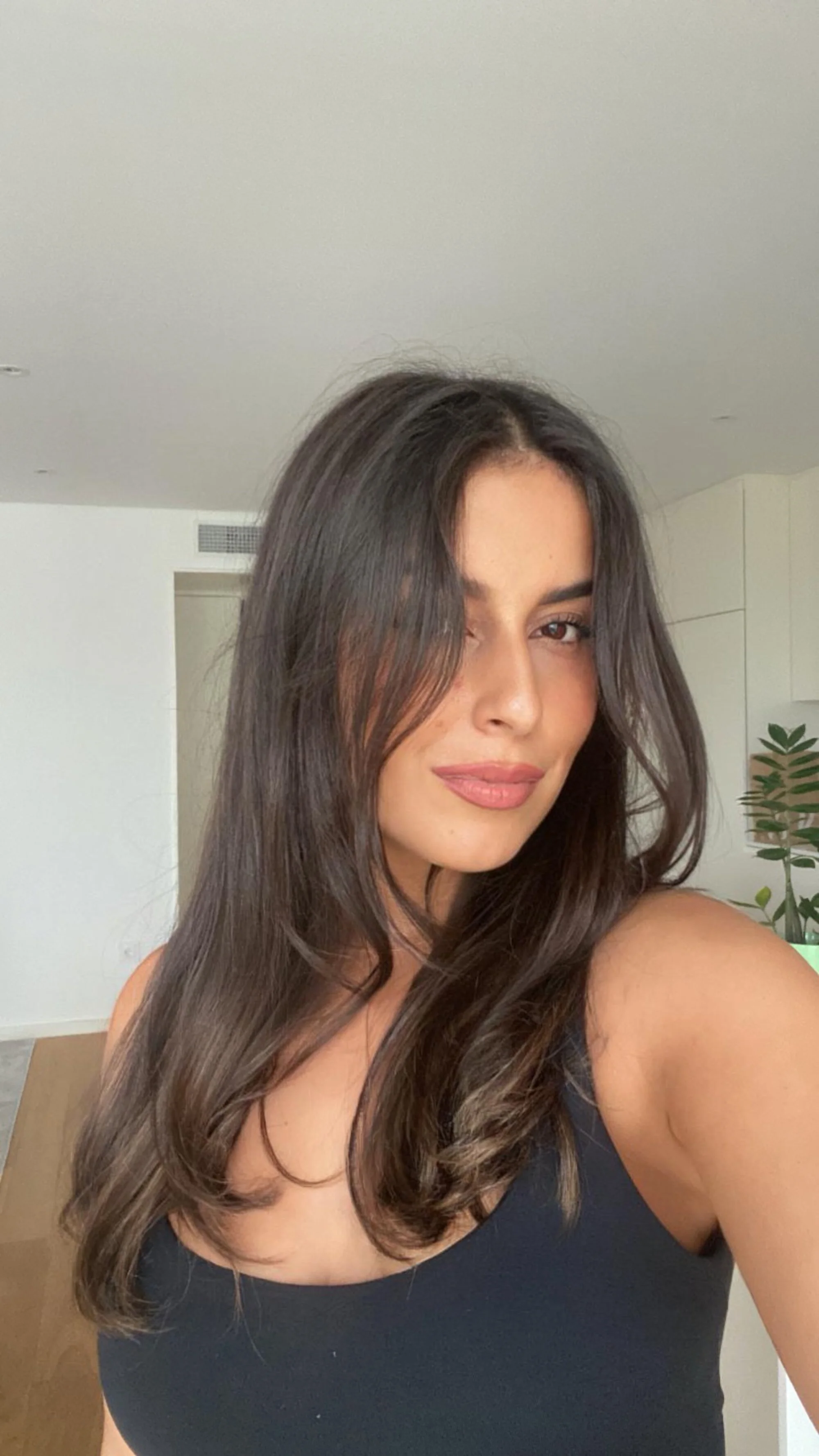 A woman with long, wavy brown hair taking a selfie indoors, wearing a black sleeveless top.
