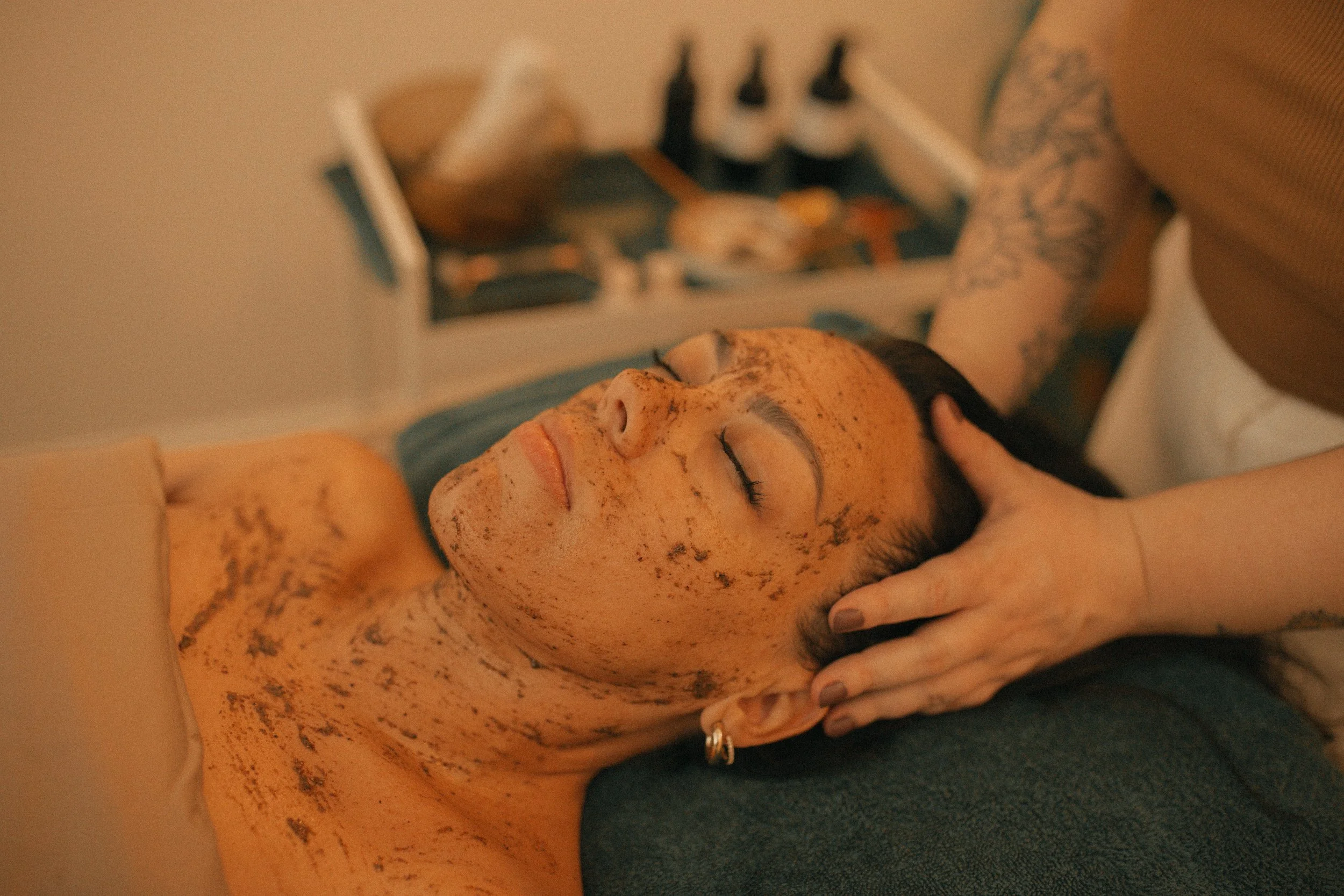 A woman with freckles and dark hair is lying on her back with her eyes closed, receiving a facial massage from a practitioner. The woman has her head resting on a pillow, and the practitioner is using both hands to massage her temples. In the background, there is a tray with bottles and skincare products.