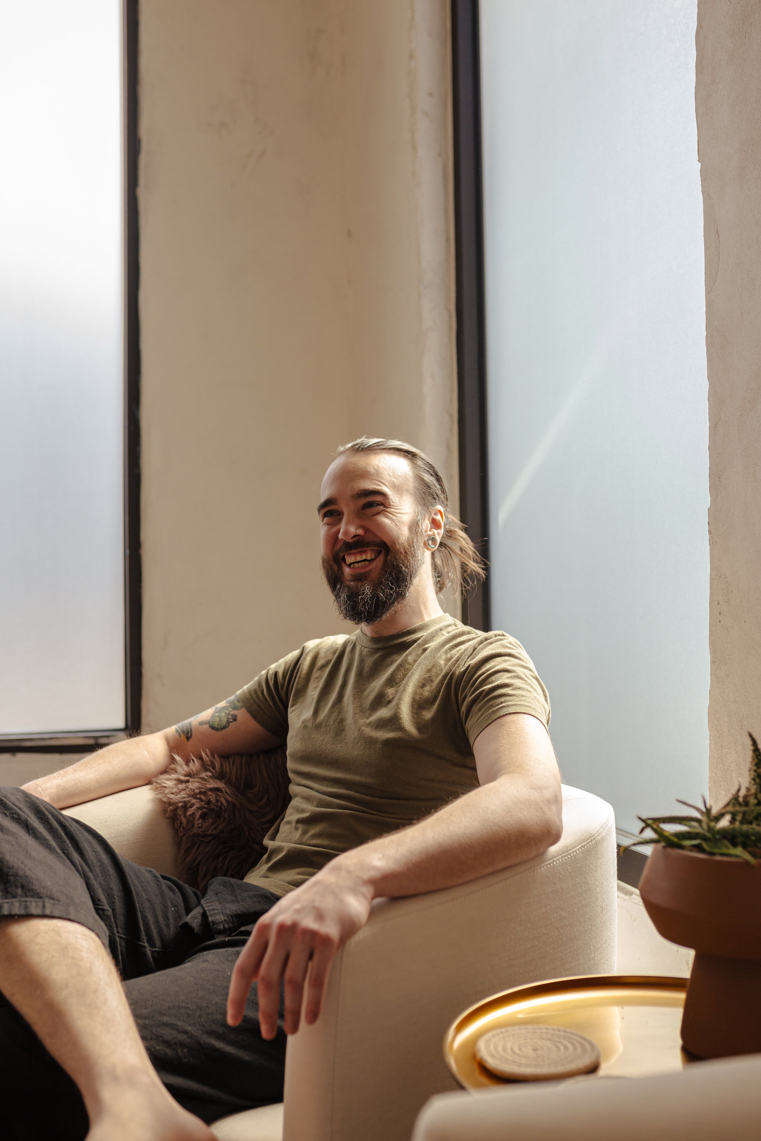 A man with a beard and long hair tied back, wearing a brown T-shirt, is sitting on a cream-colored armchair, smiling and laughing in a bright room with large windows.