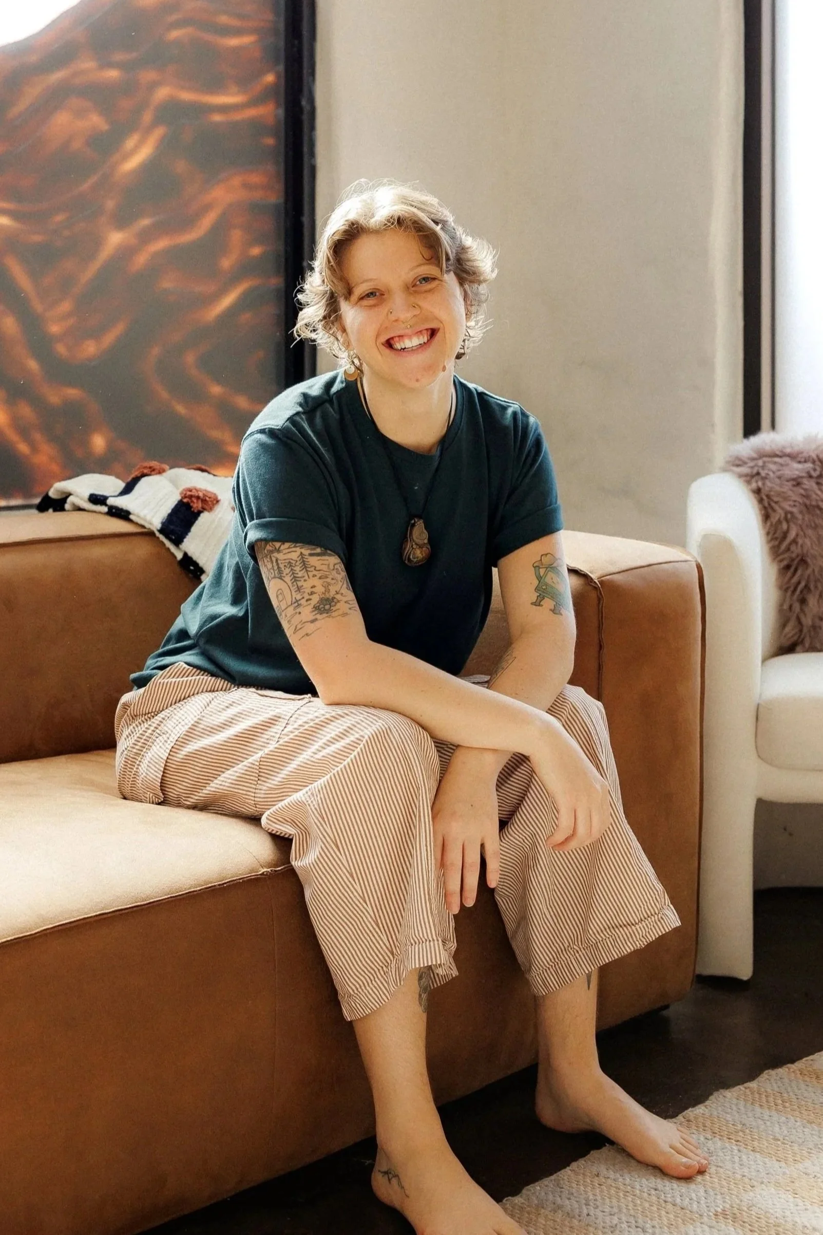 A woman with short, curly hair sitting on a white couch near large frosted windows, wearing a beige crocheted top and patterned shorts, with tattoos on her arms and earrings.