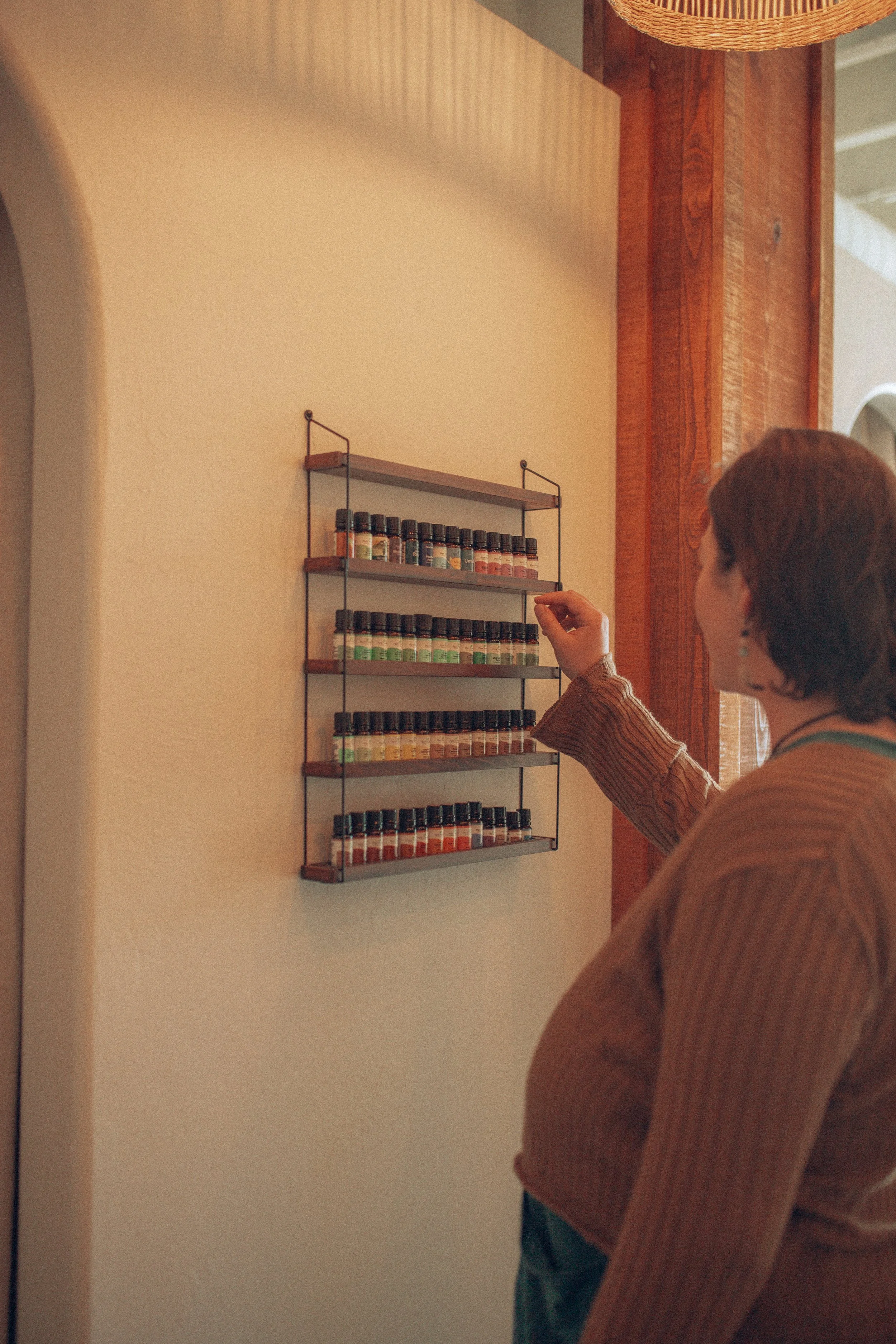 A woman wearing a striped shirt sorting essential oils on a wall-mounted shelf in a cozy, rustic room.