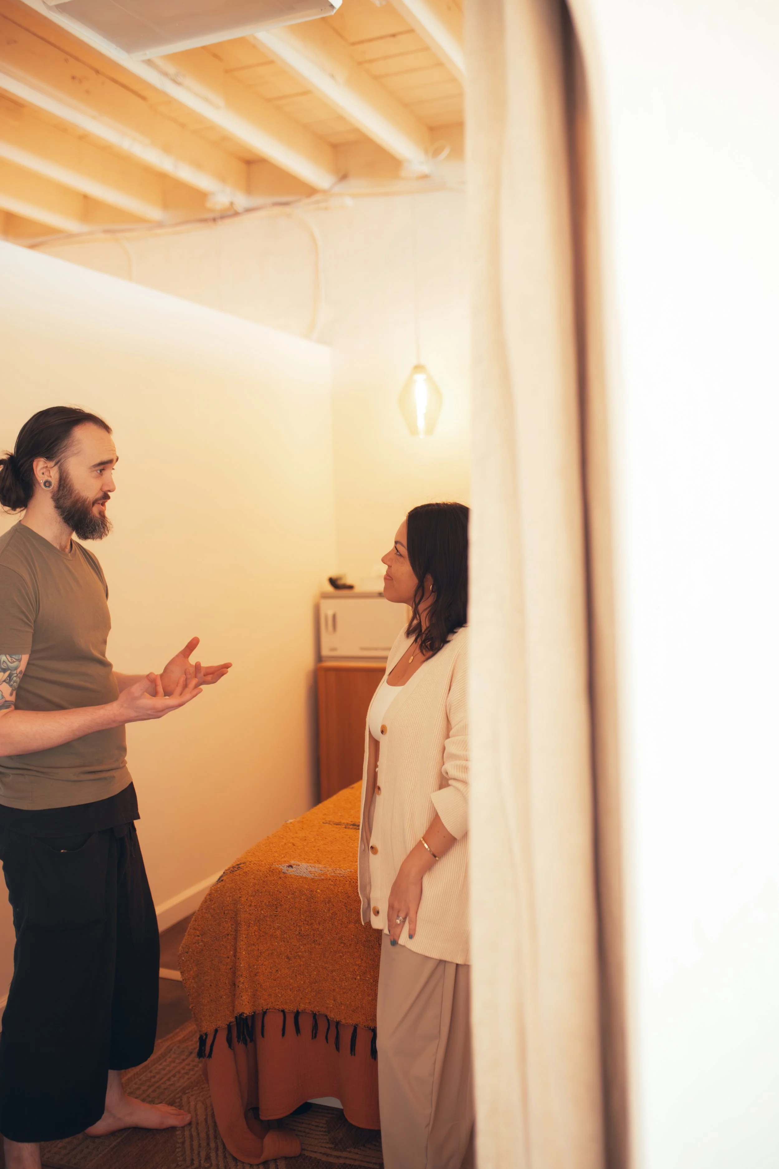 A man with a beard and tattoos, wearing a green t-shirt, is talking to a woman with black hair, wearing a white cardigan, in a room with a wooden ceiling and hanging light. The photo is taken from behind a curtain or a door.