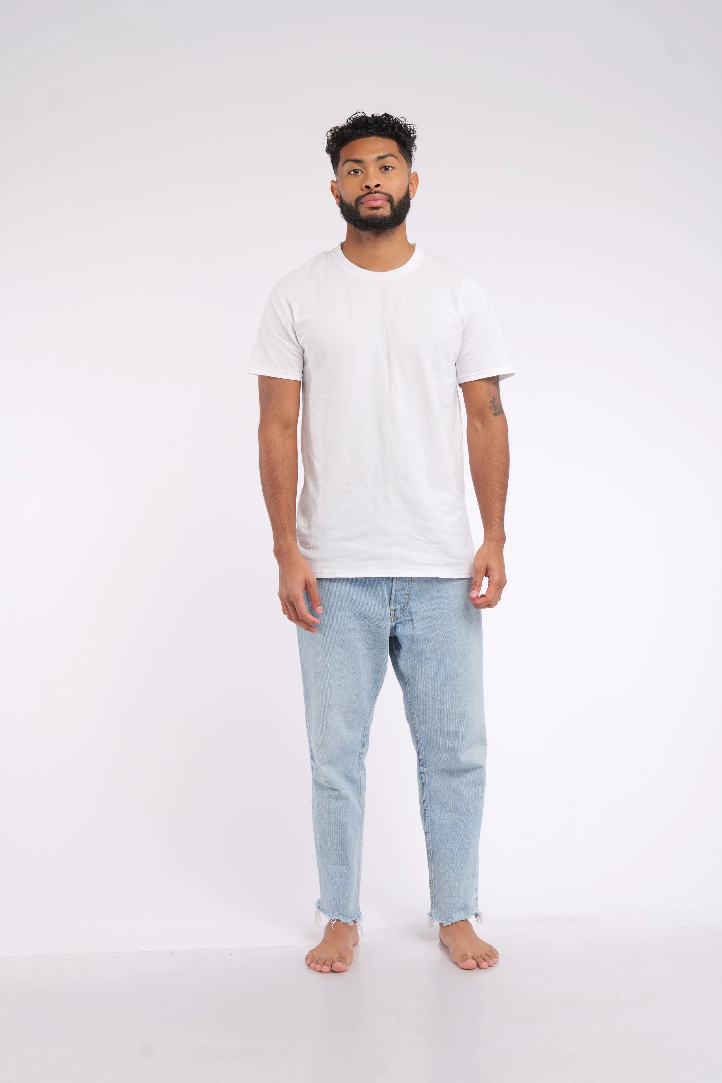 Young man standing barefoot against white background, wearing white t-shirt and light blue jeans.