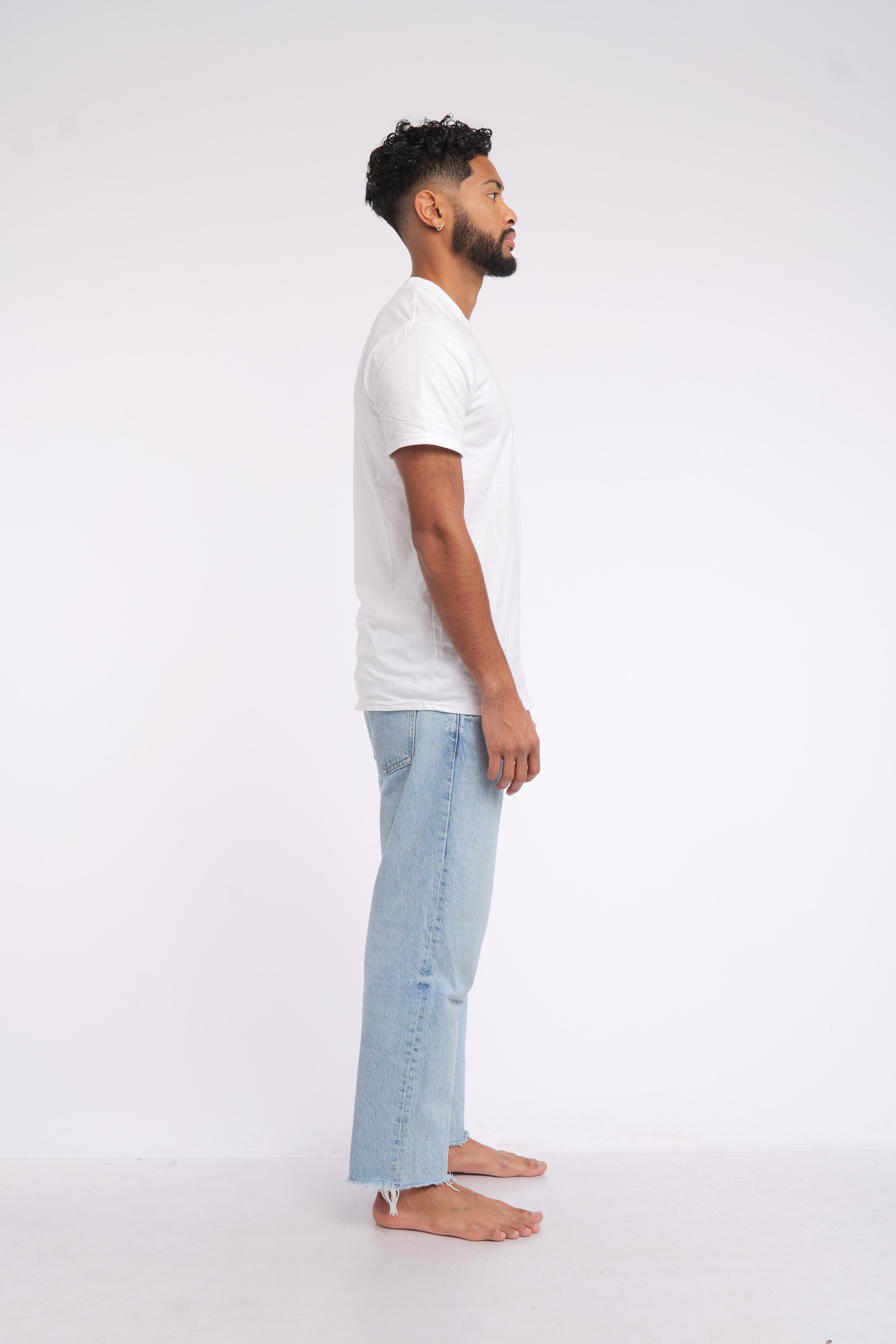 Side view of a man with dark curly hair and a beard, standing barefoot against a white background, wearing a plain white t-shirt and light blue jeans.